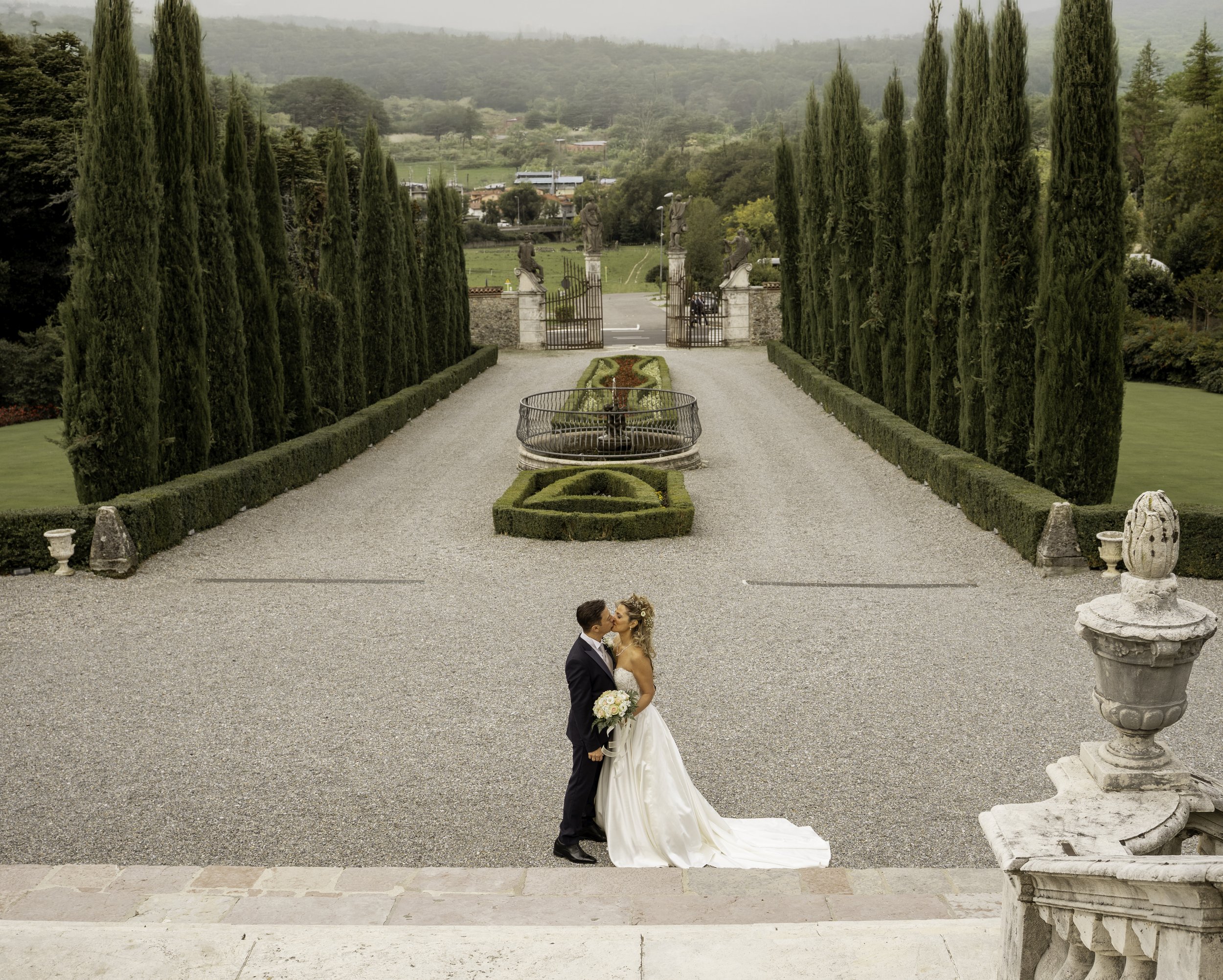 A bride and groom kissing outdoors near stone columns and a decorative garden centerpiece.