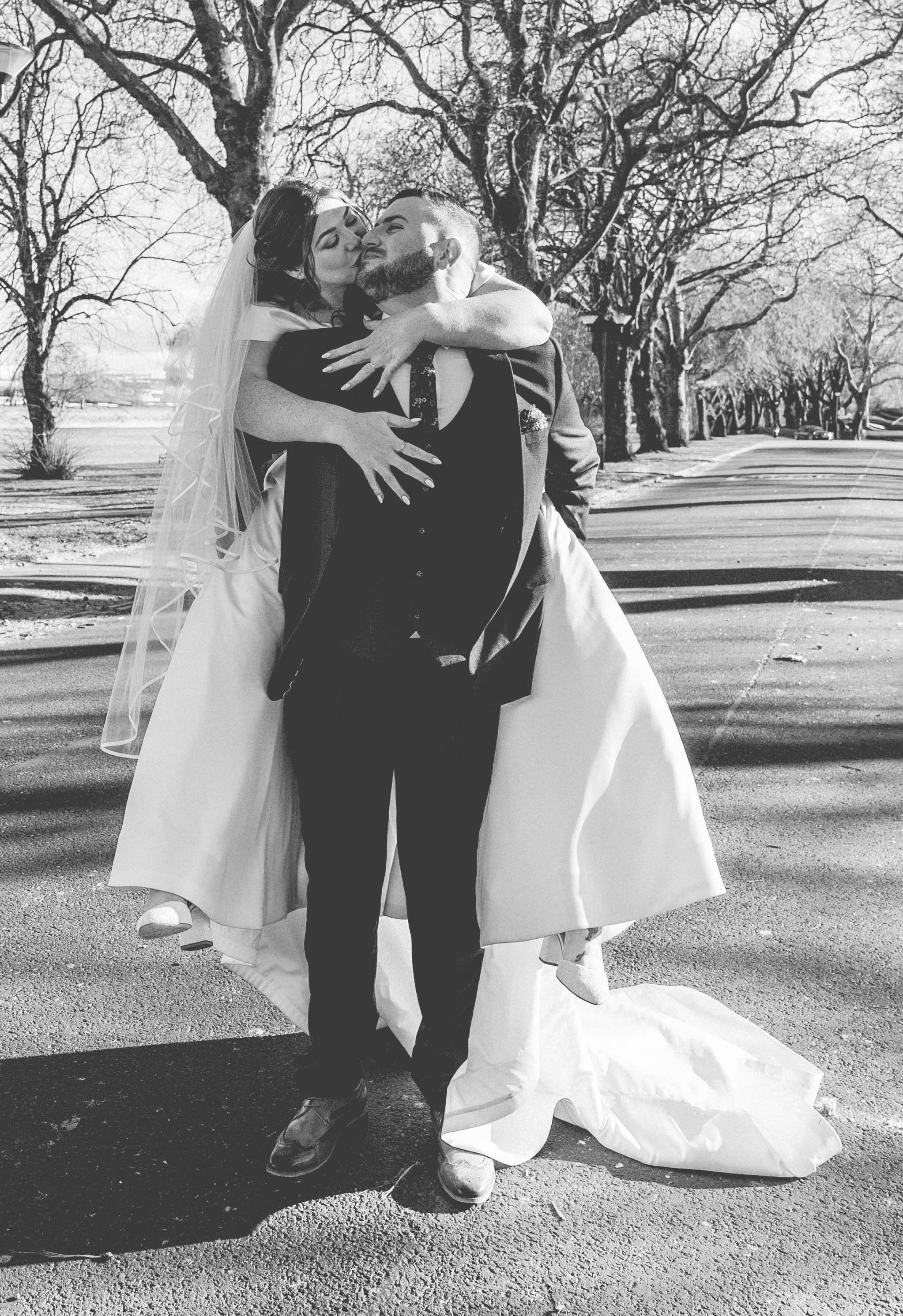 Black and white photo of a newlywed couple embracing outdoors, with trees and a paved road in the background. The bride is wearing a wedding dress with a veil, and the groom is dressed in a suit.