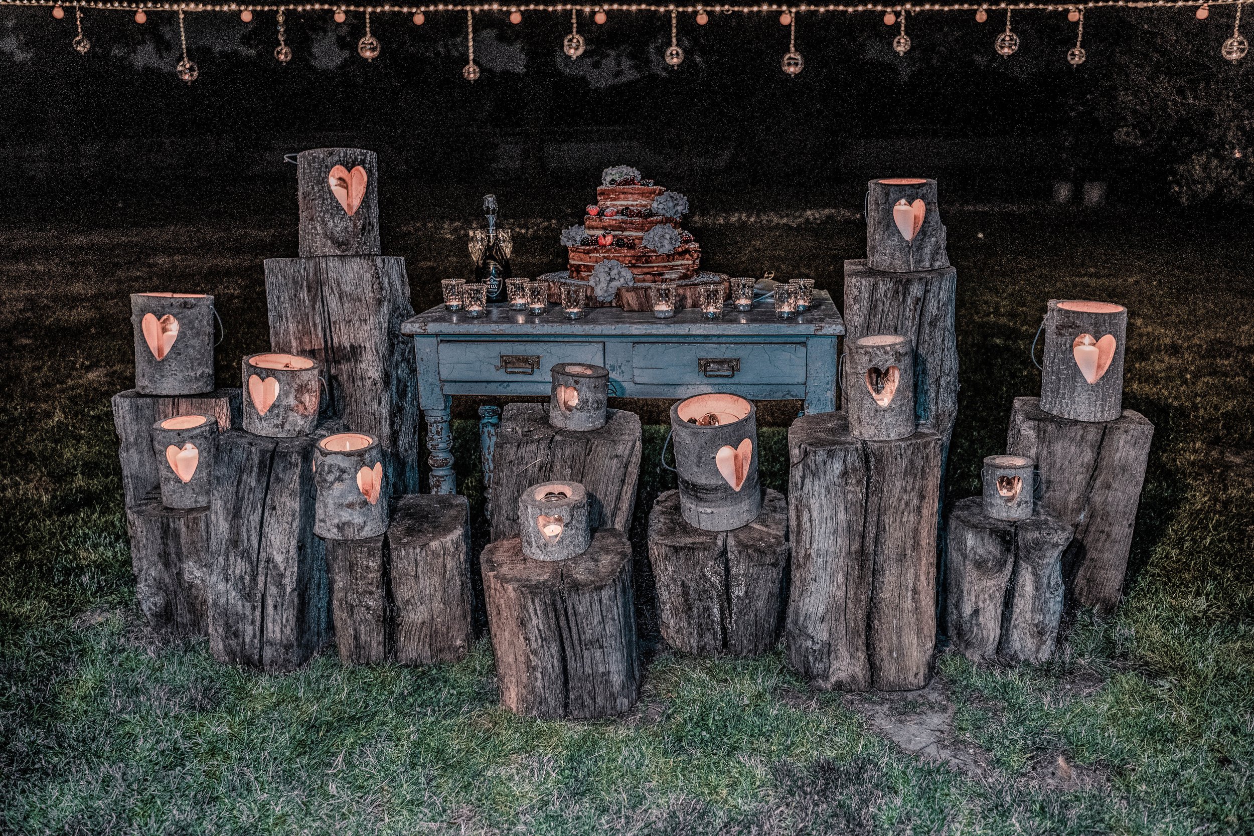 Outdoor wedding or romantic celebration setup with rustic wooden logs arranged in a semi-circle, each with a lit candle inside. A blue vintage table in the center holds a three-tiered cake decorated with flowers, along with a small bottle and glasses
