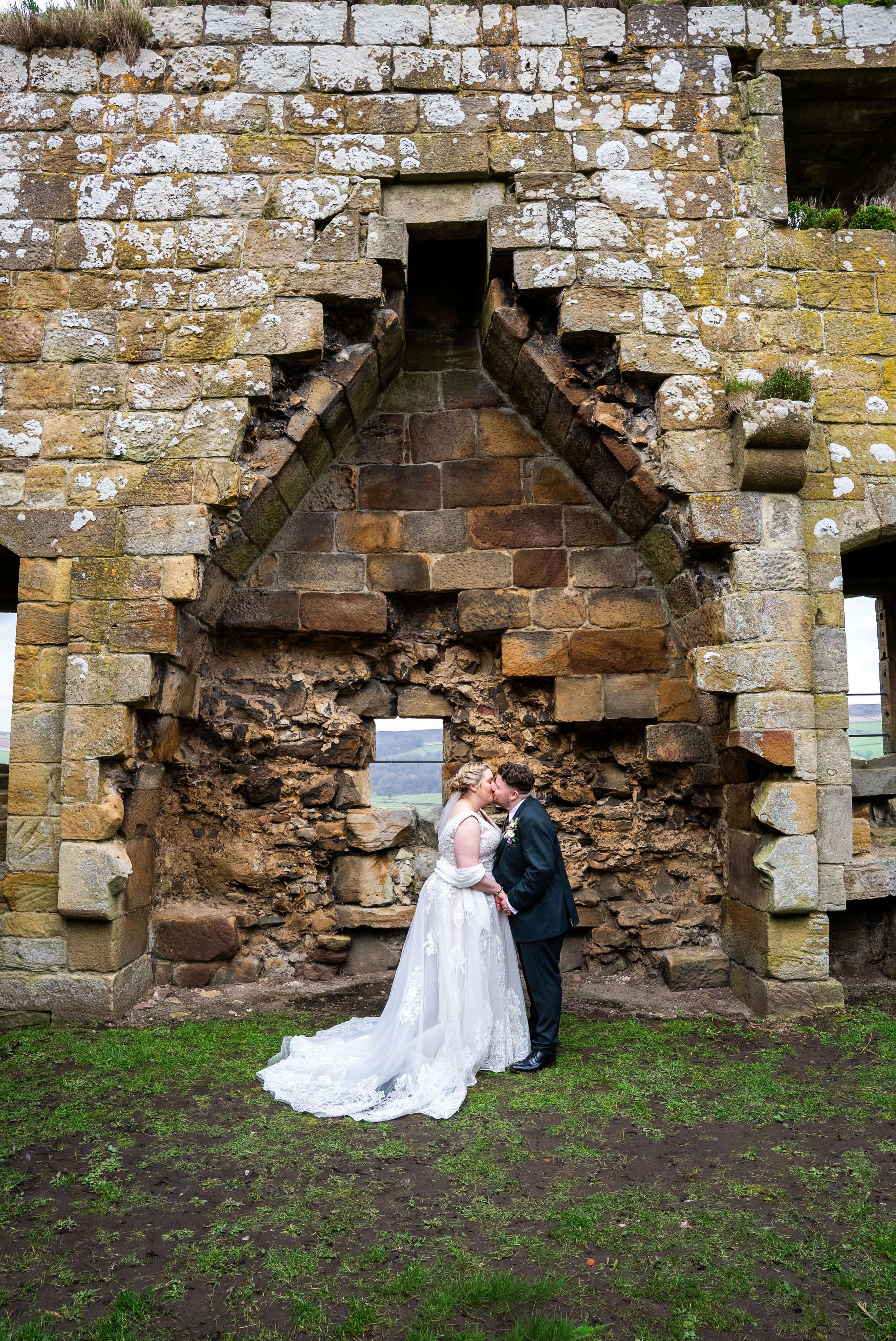 Bride and groom kiss in front of a historic stone structure with a large alien-like arch opening.