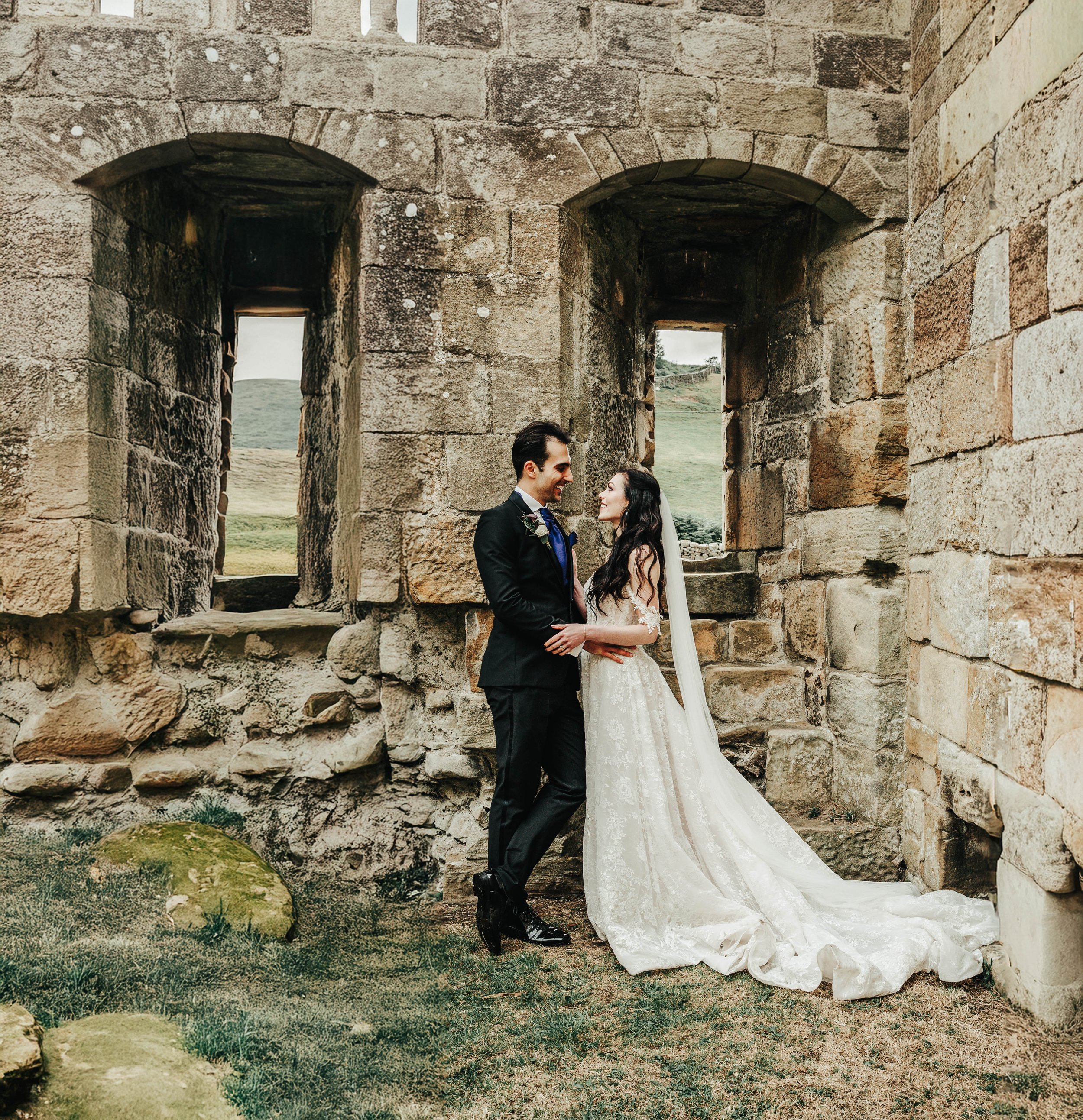 A bride and groom standing close together outdoors near ancient stone ruins, looking at each other and smiling, with hills and cloudy sky in the background.