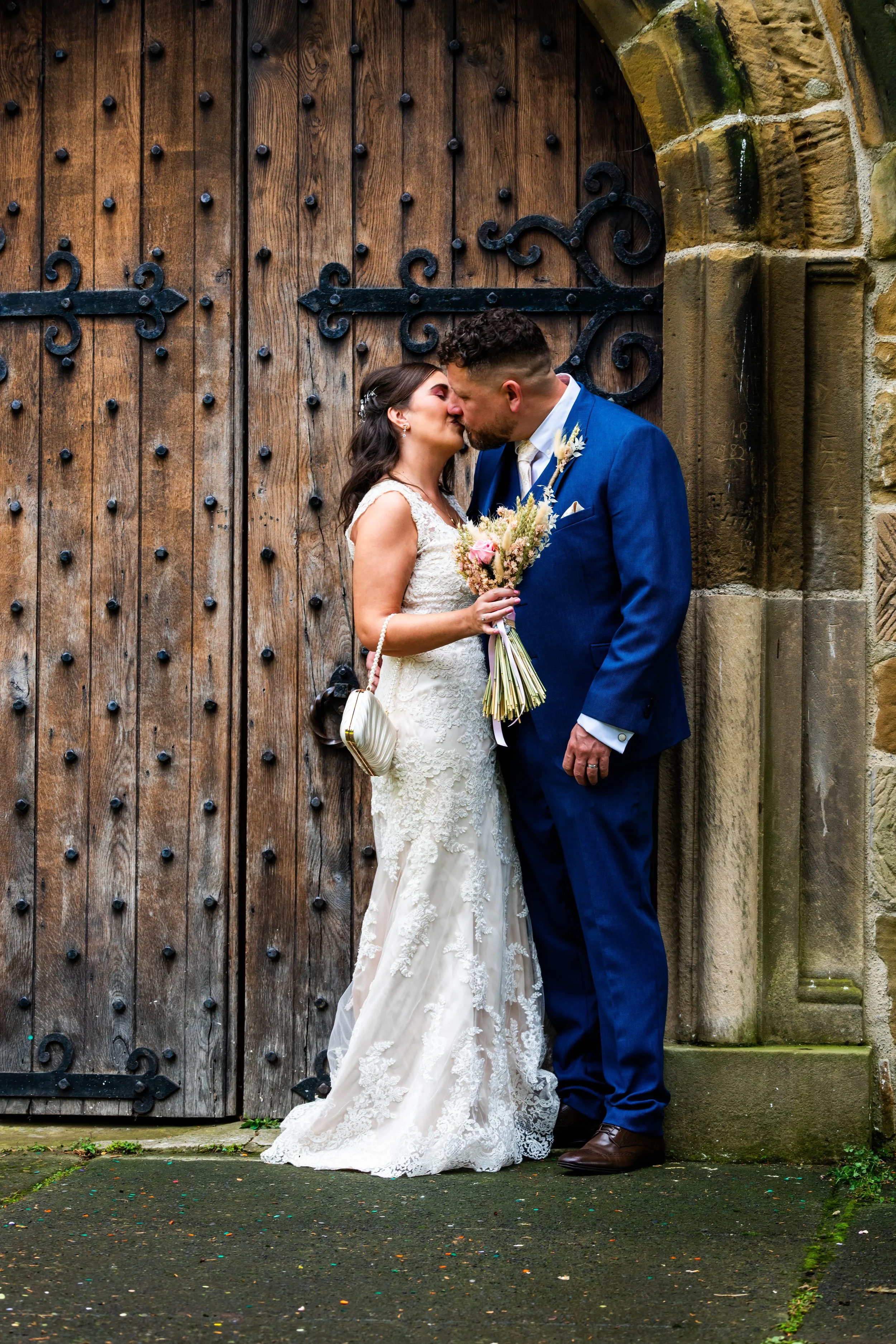 A bride and groom sharing a kiss in front of a wooden door with decorative black iron hinges, on a stone and moss-covered ground.