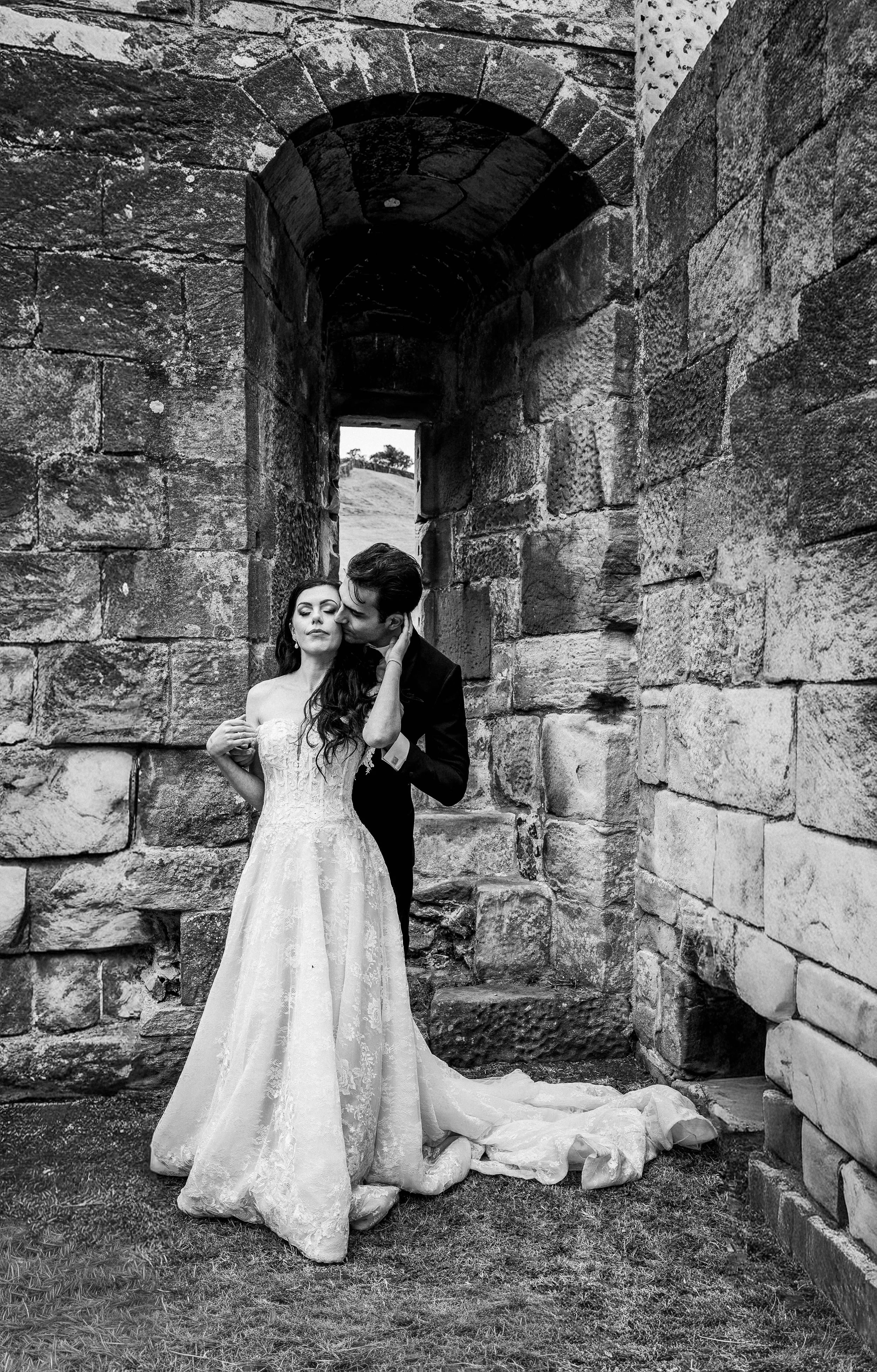 Black and white photo of a bride and groom in wedding attire, standing close together under a stone archway at an outdoor location, with the groom kissing the bride's cheek.