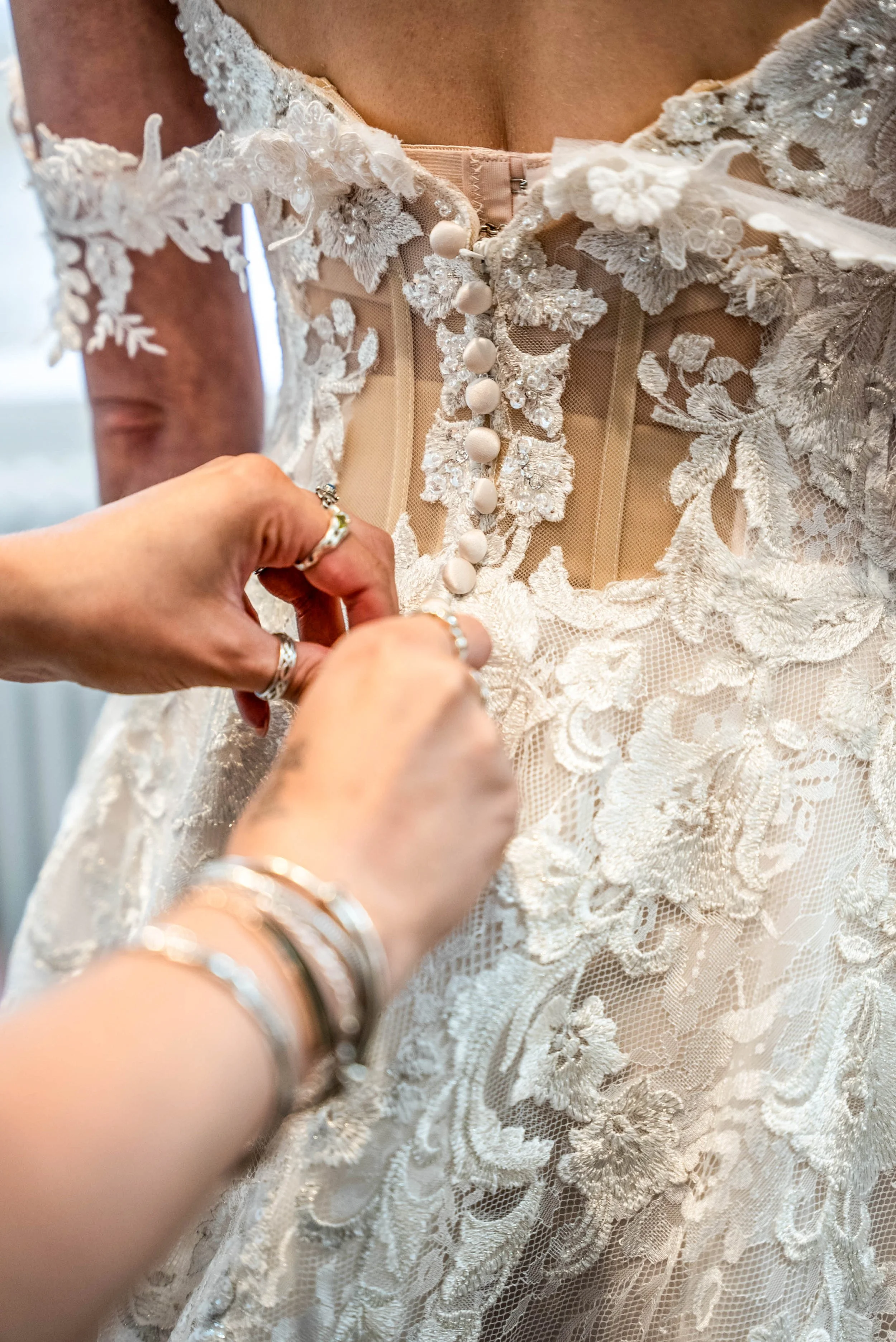 Close-up of a bride's dress with a person fastening the row of buttons on the back, featuring intricate lace and floral detailing.