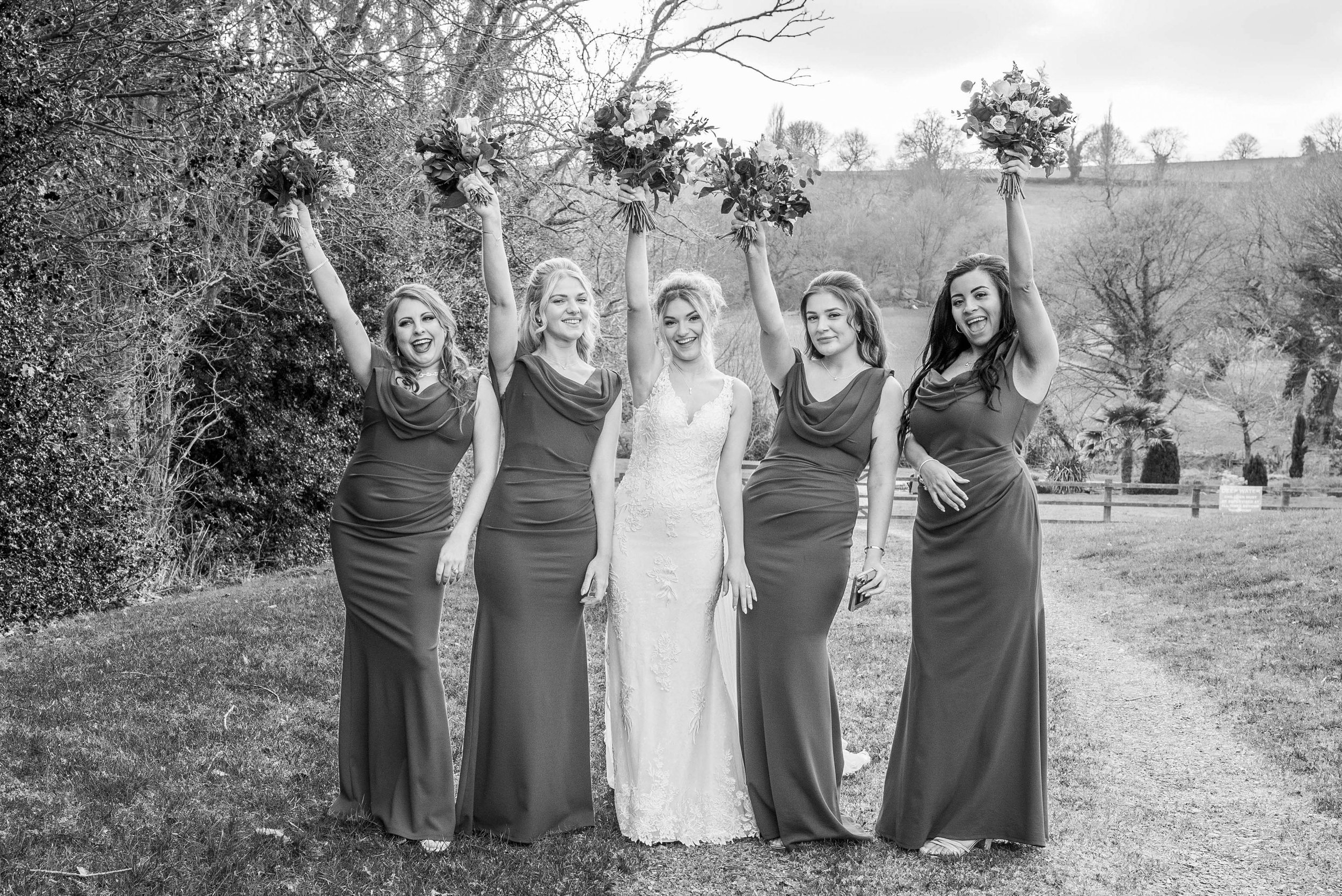 Black and white photo of five women outdoors, four bridesmaids and one bride, celebrating with the bridesmaids holding bouquets raised in the air.