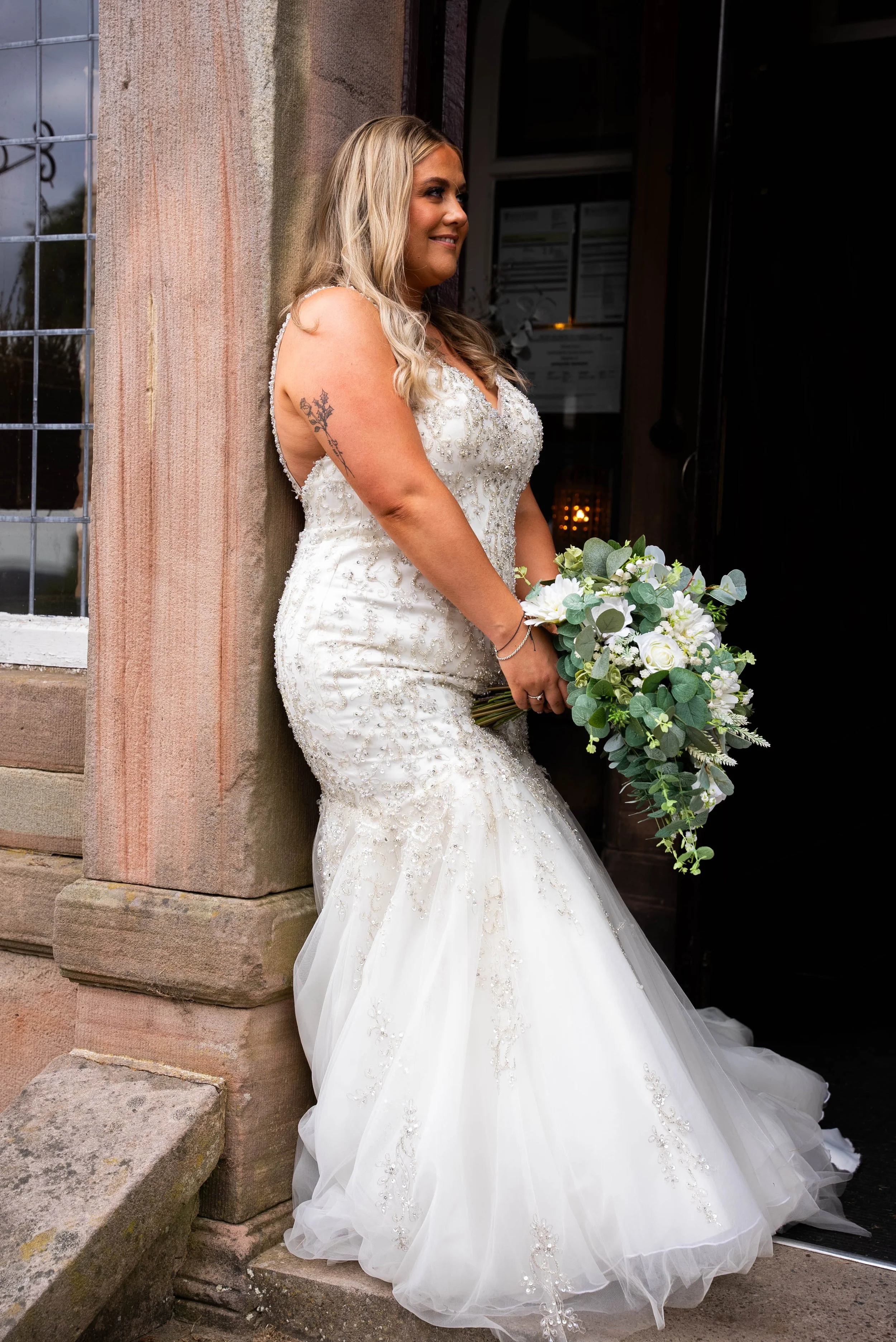 A bride in a white, embellished wedding gown holding a bouquet of white roses and greenery, leaning against a stone building wall outside a venue.
