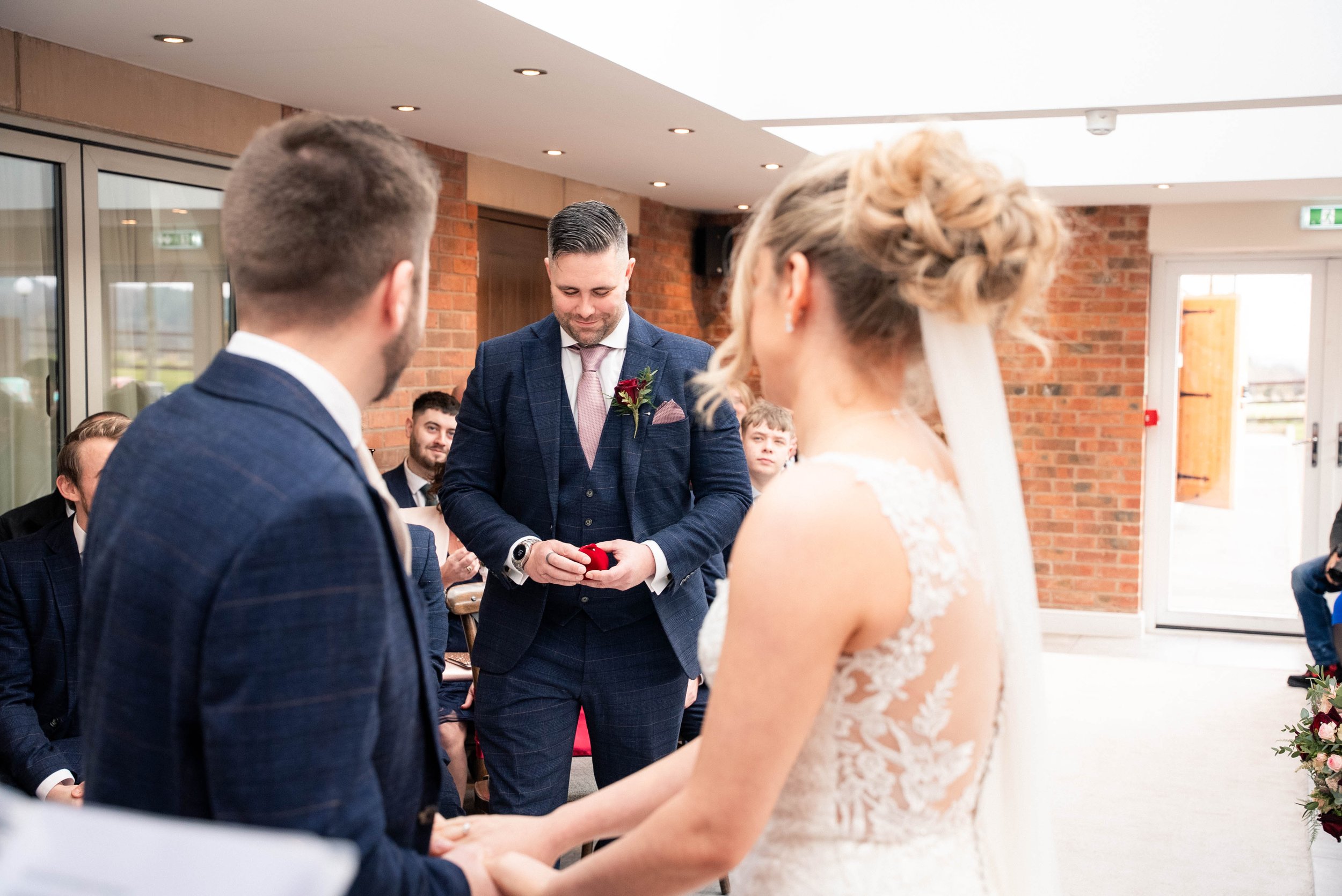A wedding ceremony where the bride and groom are holding hands, facing each other, with a man in a blue suit officiating. Guests are seated in the background inside a room with brick walls and large glass doors.