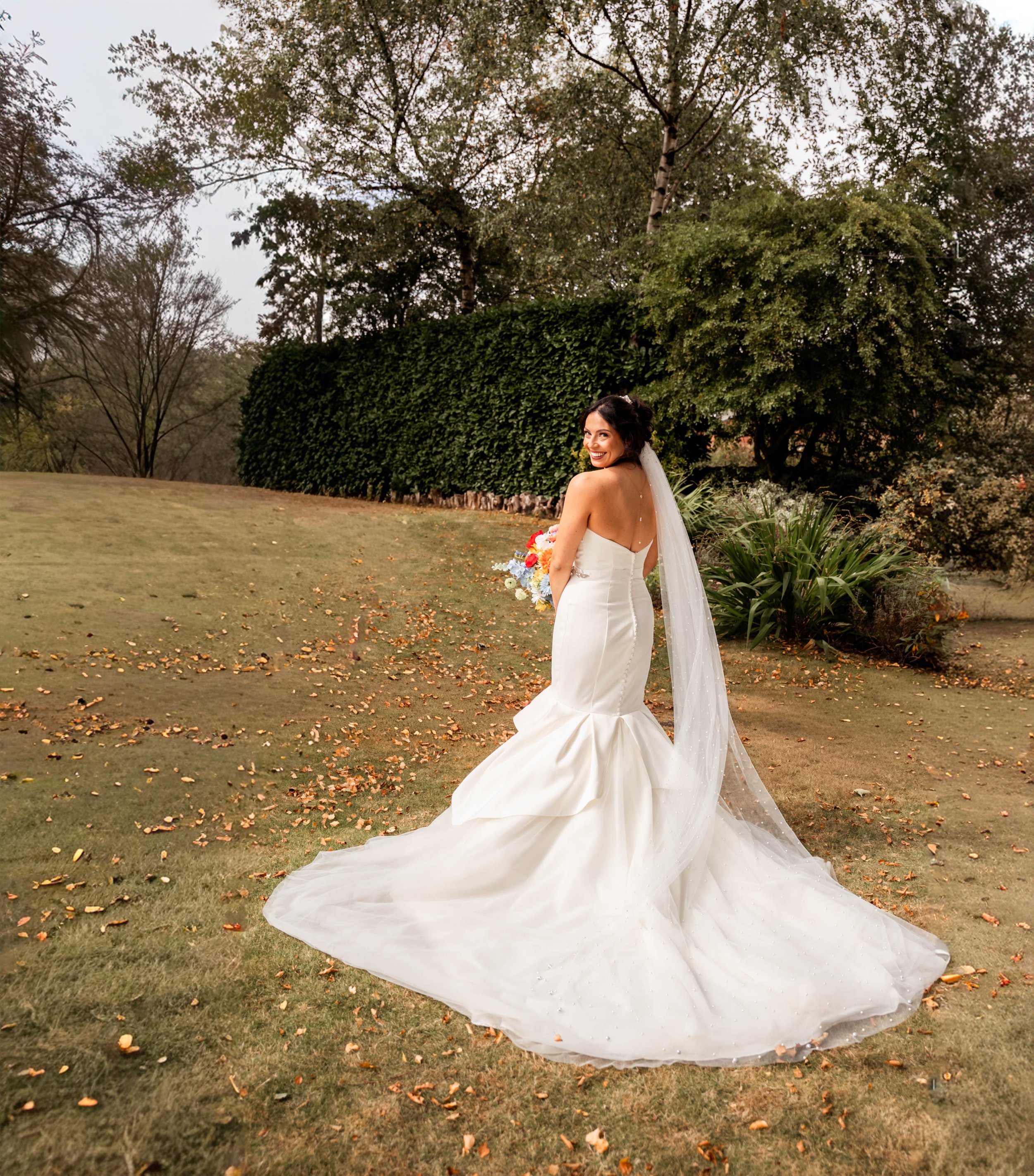 Bride in a white wedding gown holding a bouquet, standing outdoors on a grassy area with trees and shrubs in the background.