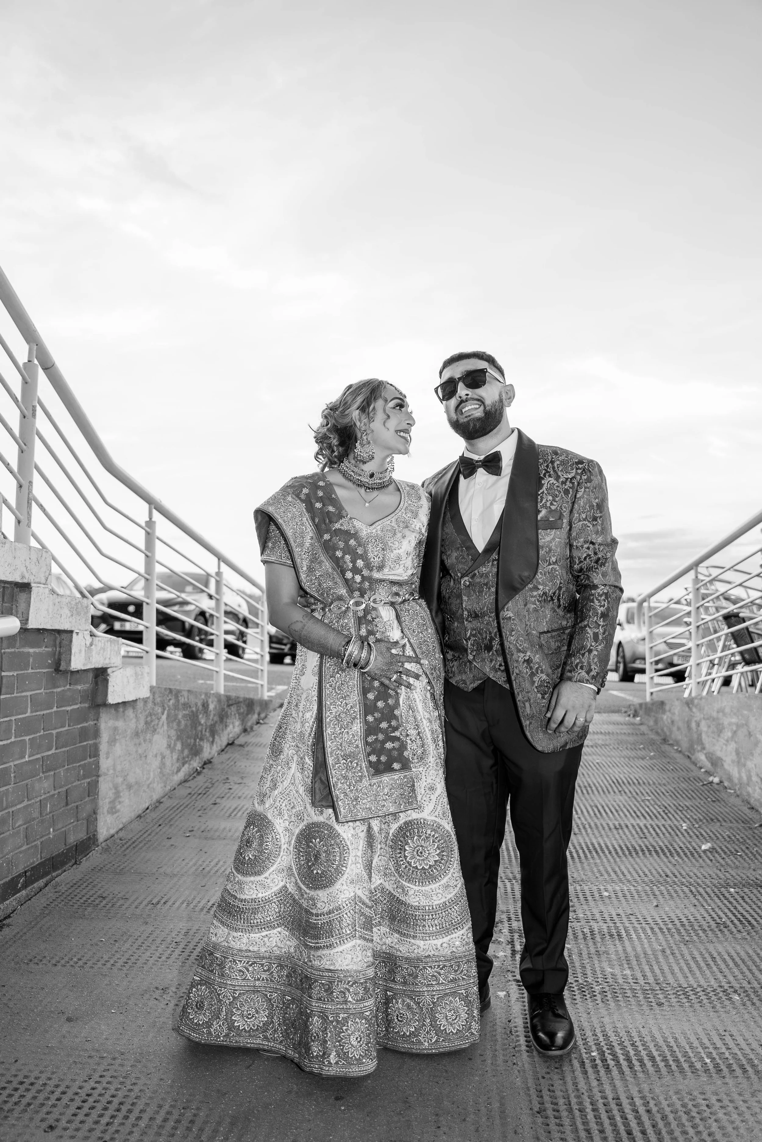 A couple dressed in formal attire standing on a ramp outdoors, smiling and looking at each other, with a parking lot in the background.