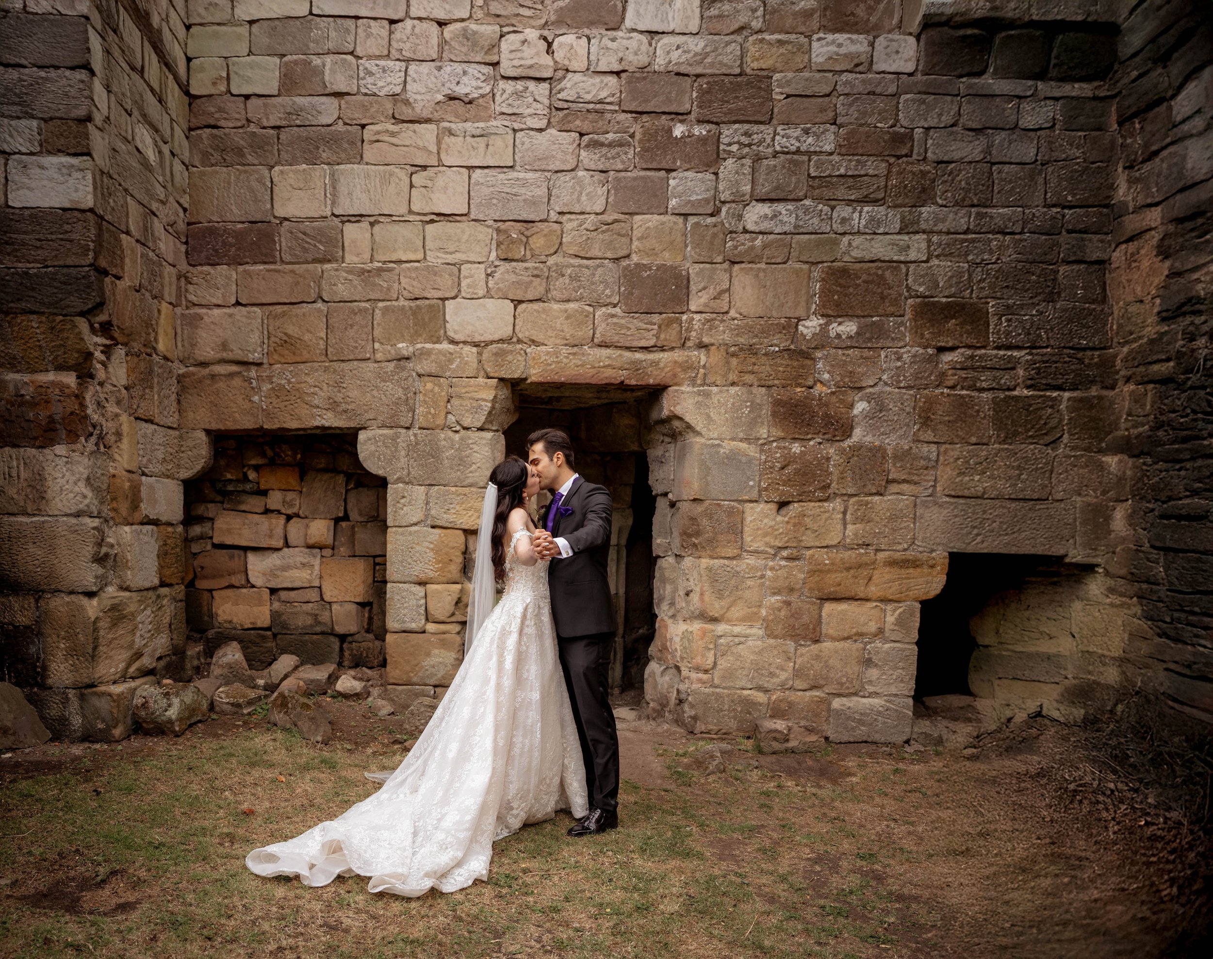 A bride and groom share a kiss in front of a rustic stone wall at their wedding.