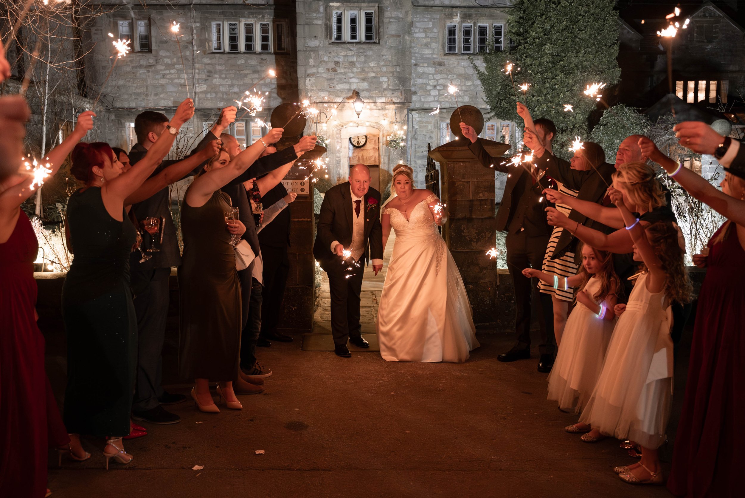 Wedding celebration outside with guests holding sparklers, bride and groom in formal attire, historic stone building in background, night scene with festive lighting.
