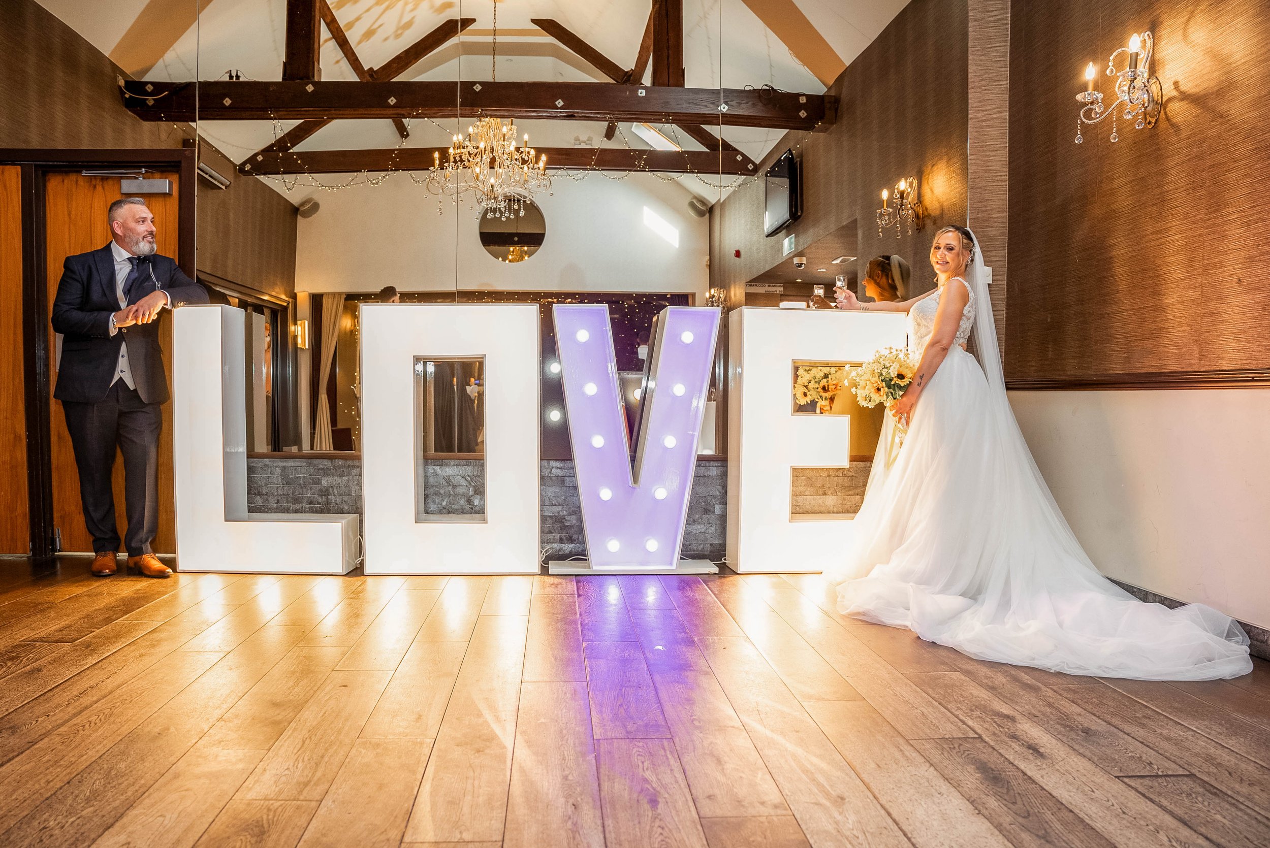 A wedding reception scene featuring a bride in a white wedding gown holding a bouquet and smiling, a groom in a dark suit leaning on a ledge, and large illuminated letters spelling 'LOVE' in the foreground.