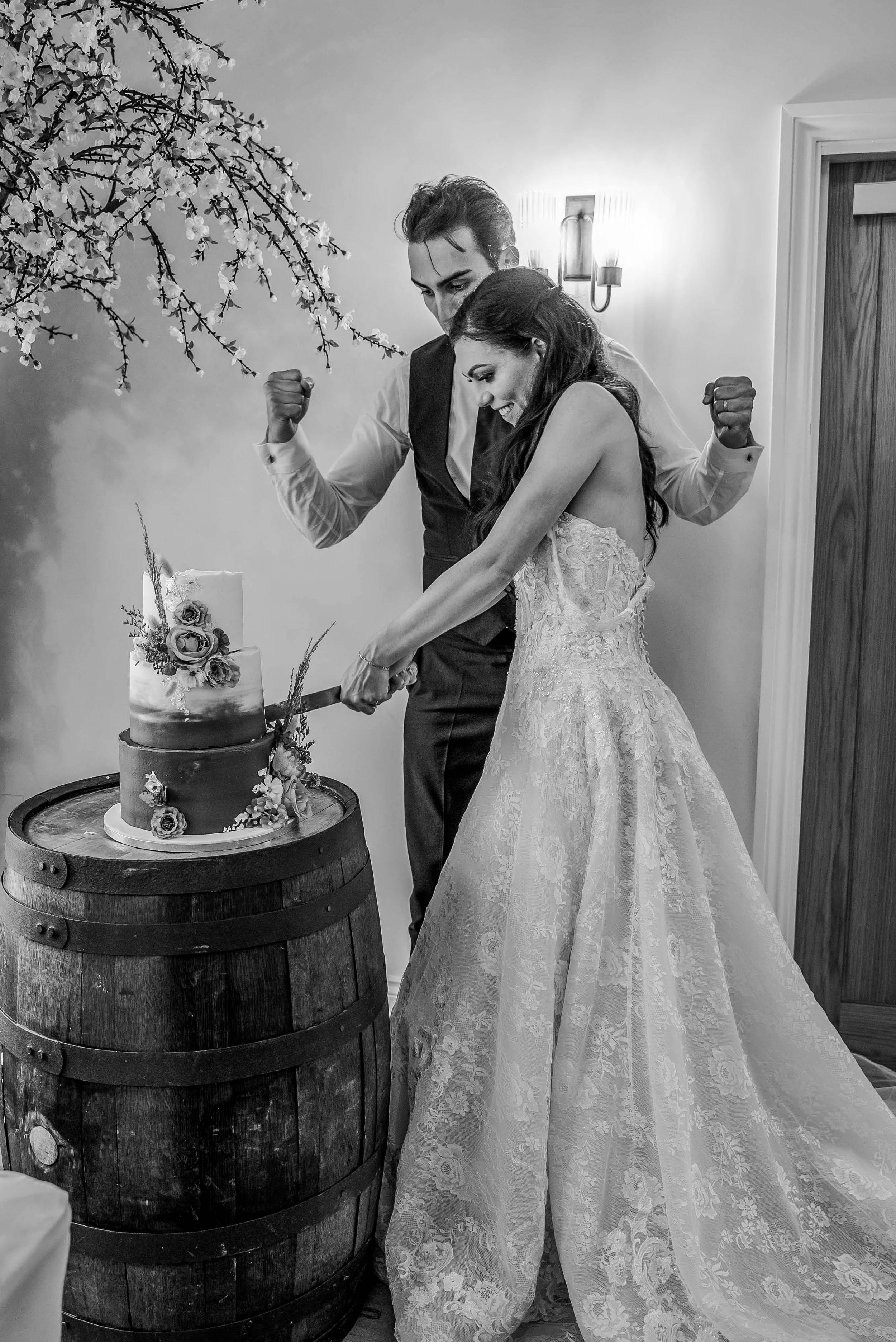 A bride and groom in wedding attire happily cutting a wedding cake together, standing behind a wooden barrel.
