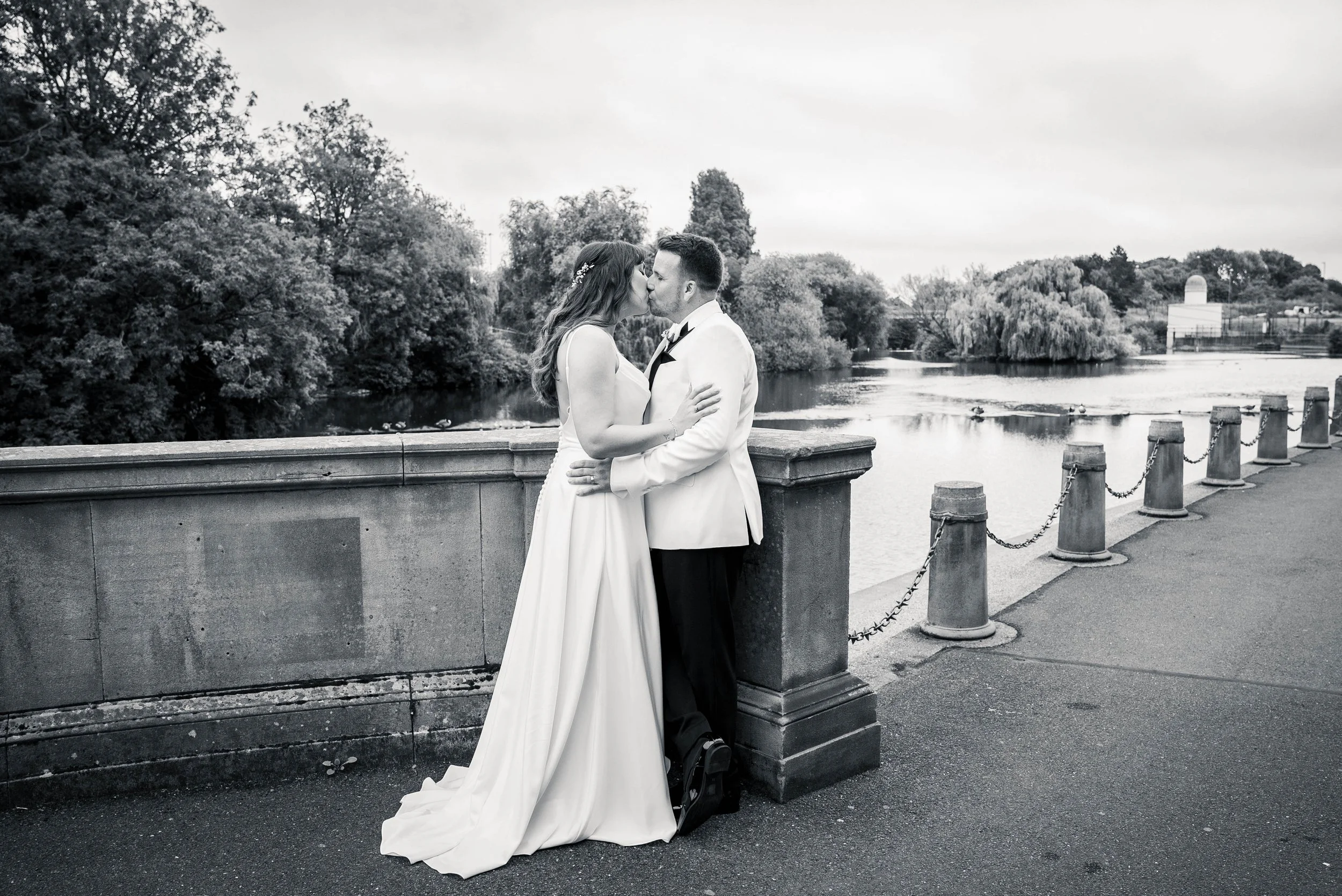 A bride and groom kissing on a bridge near a river with trees and cloudy sky in the background, in black and white.
