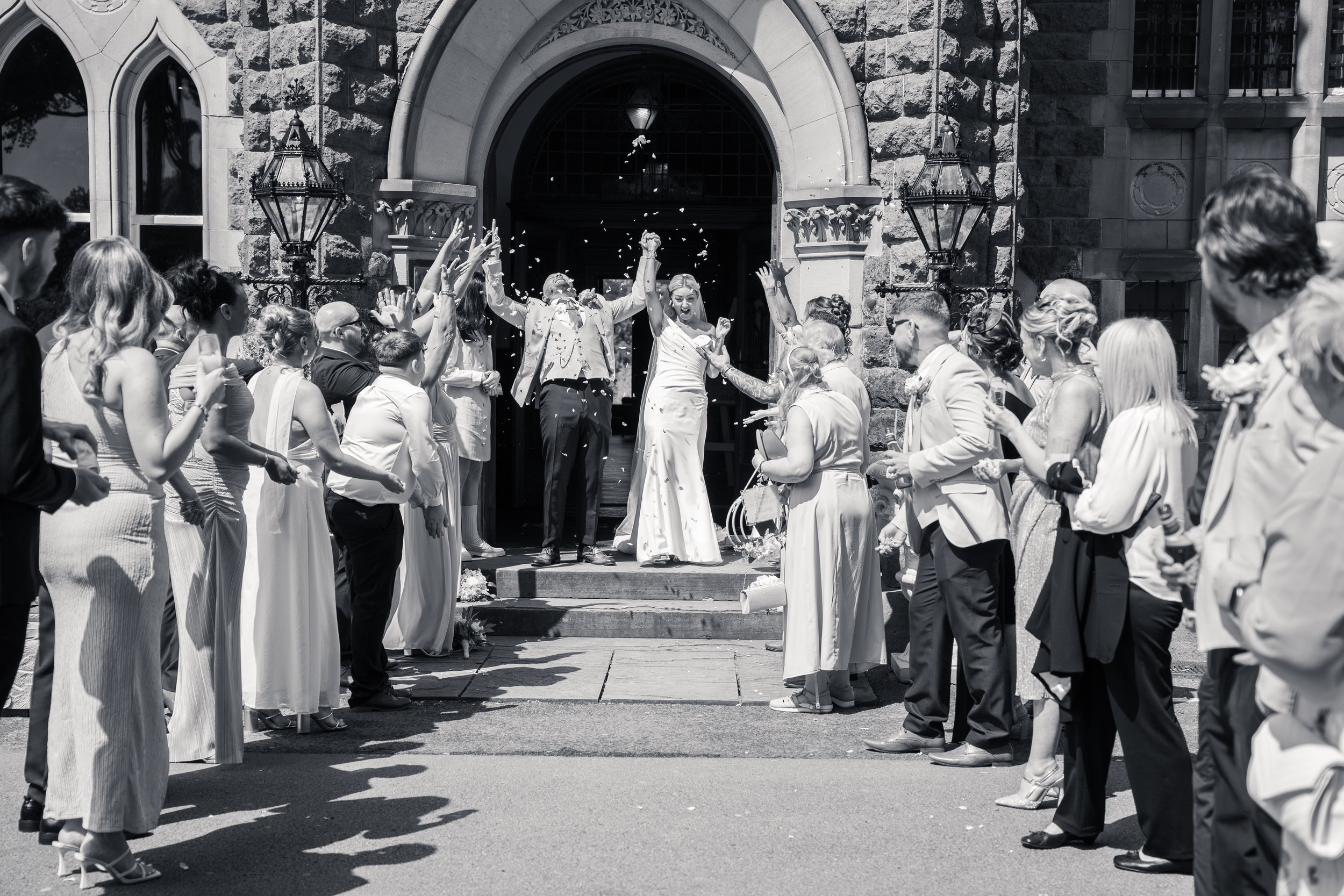A wedding celebration outside a stone church with guests forming a semi-circle, throwing rice or confetti, while the bride and groom stand at the entrance, celebrating the newlywed couple.