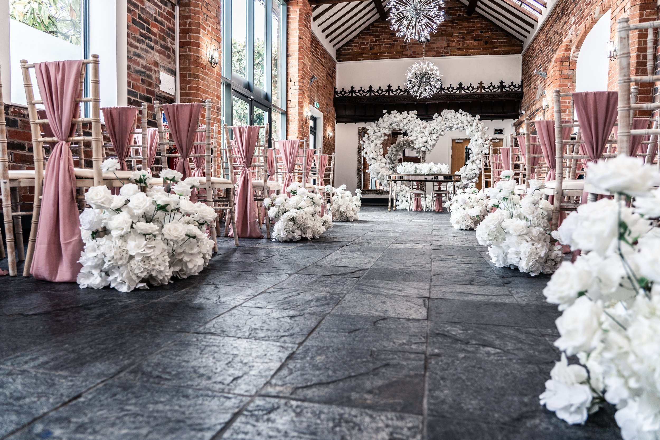 Wedding ceremony setup inside with rows of chairs draped in pink and decorated with white flowers, a floral arch at the front, brick walls, large windows, and modern chandeliers hanging from the ceiling.