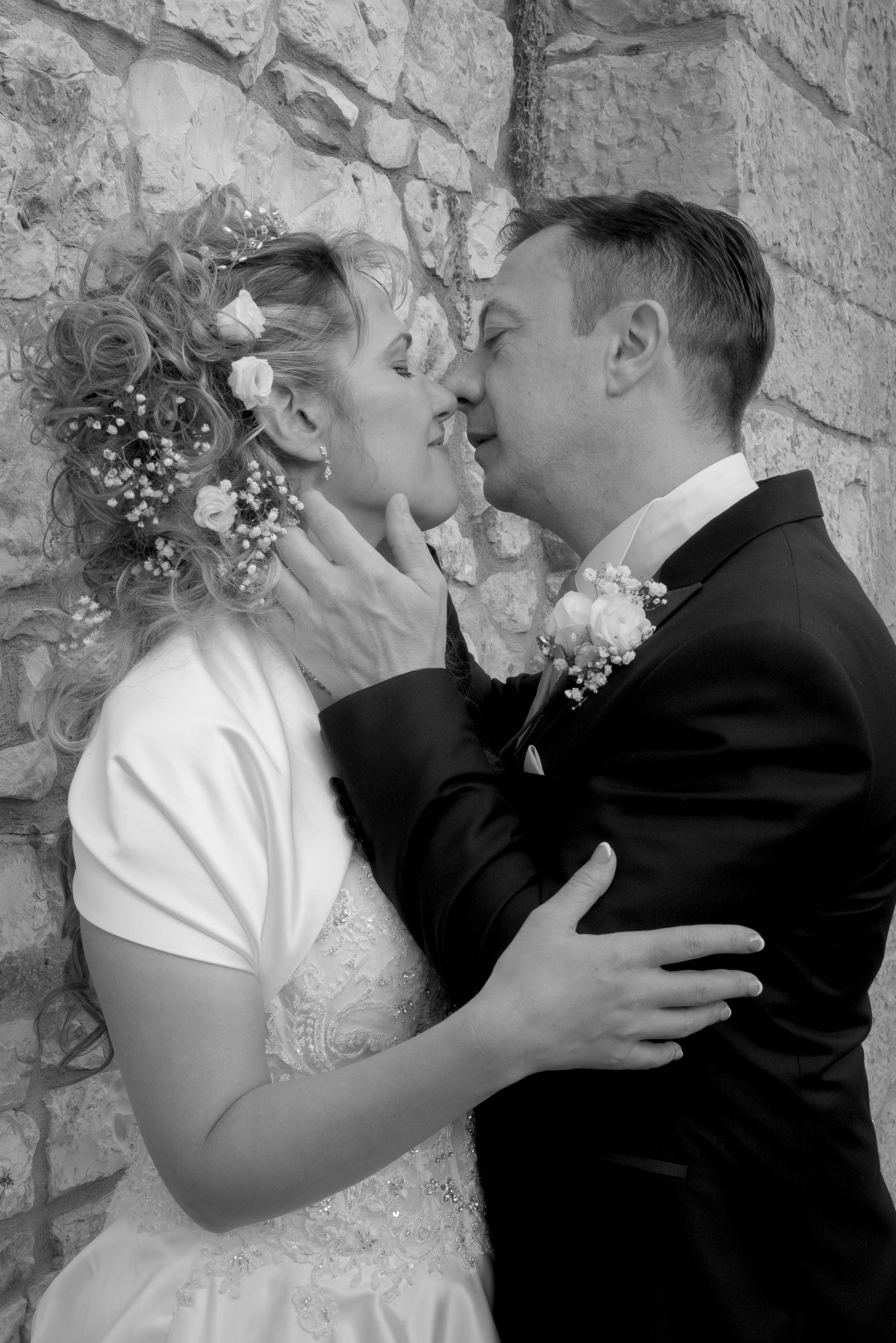 A black and white photo of a bride and groom close to each other, about to kiss. The bride has curly hair decorated with flowers and a veil, and the groom is dressed in a tuxedo with a boutonniere. They are standing in front of a stone wall, with the