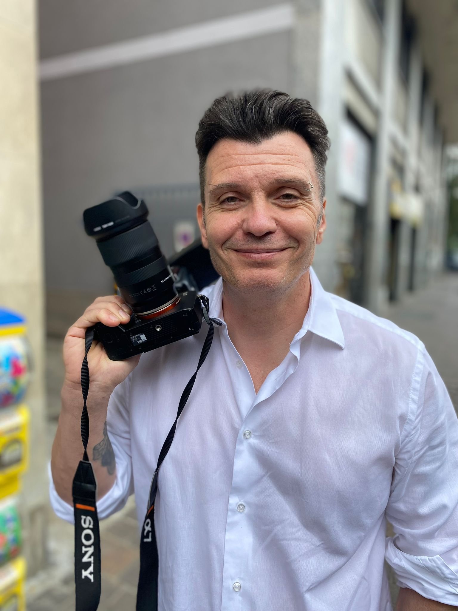 A man with dark hair and a ring eyebrow piercing, wearing a white button-up shirt, smiling and holding a Sony camera strap.