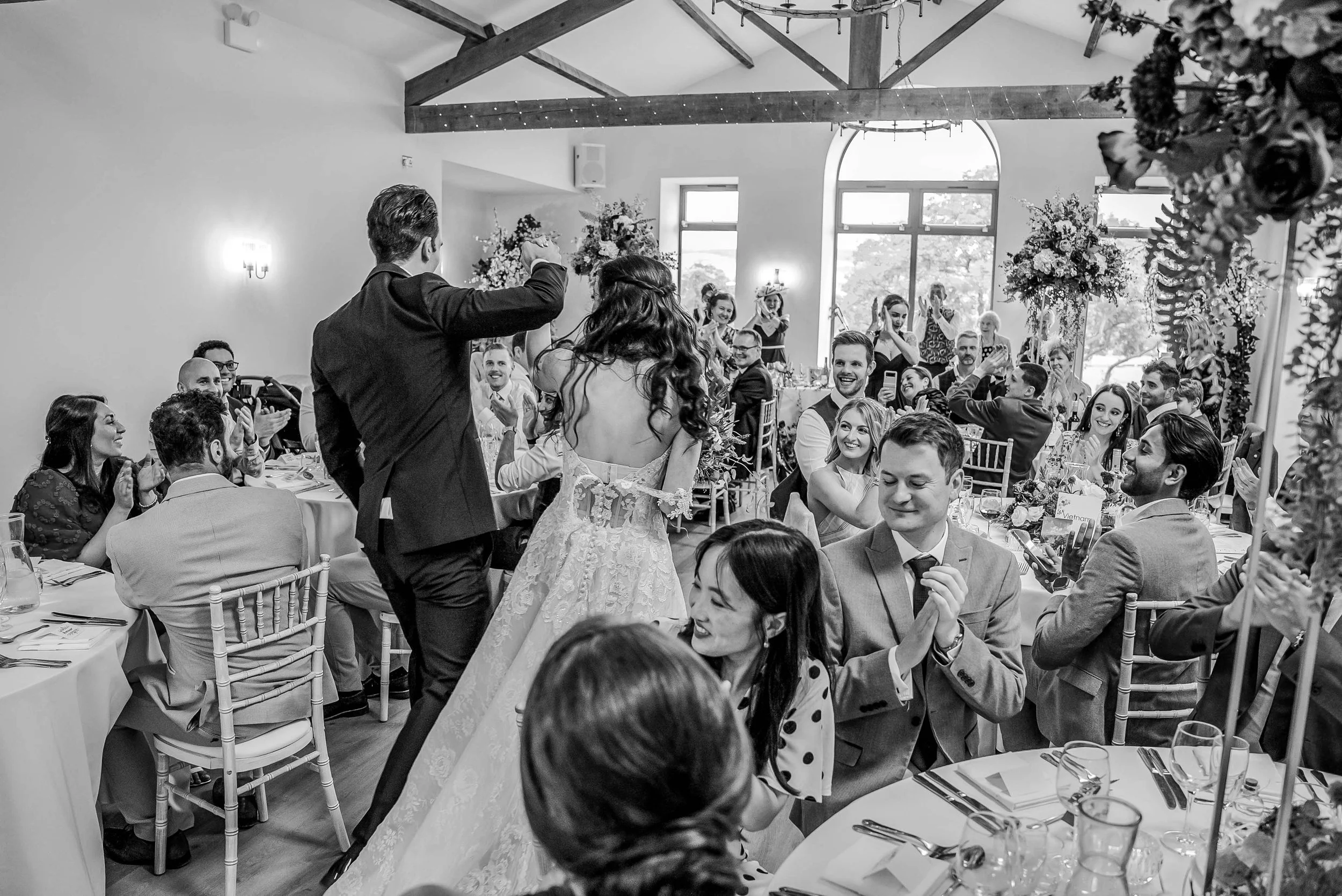 Black and white photo of a wedding reception with a bride and groom dancing among guests seated at decorated tables.
