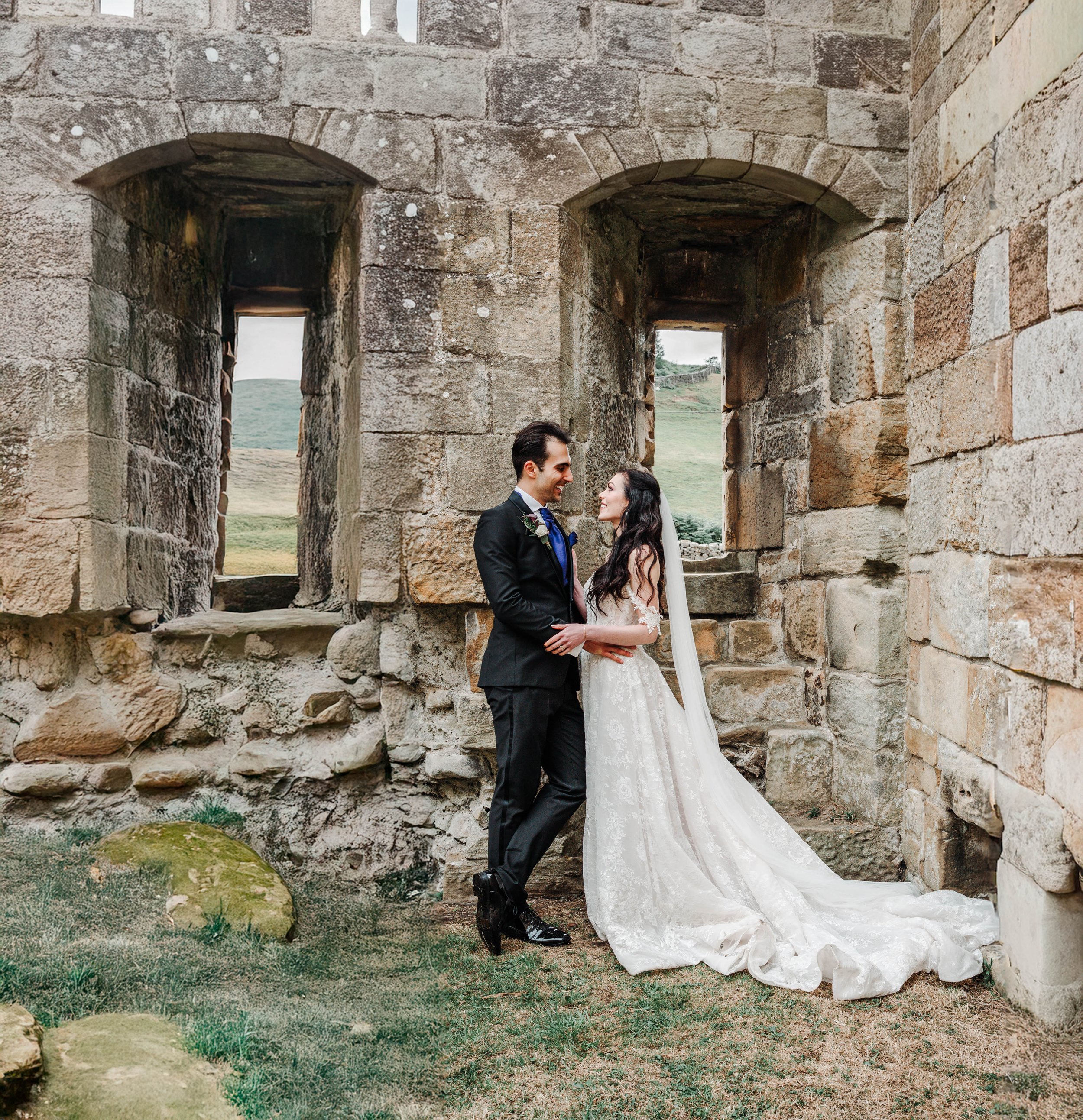 A bride and groom in wedding attire standing close together and smiling at each other in front of the stone ruin with two arched windows, with a view of green hills in the background.