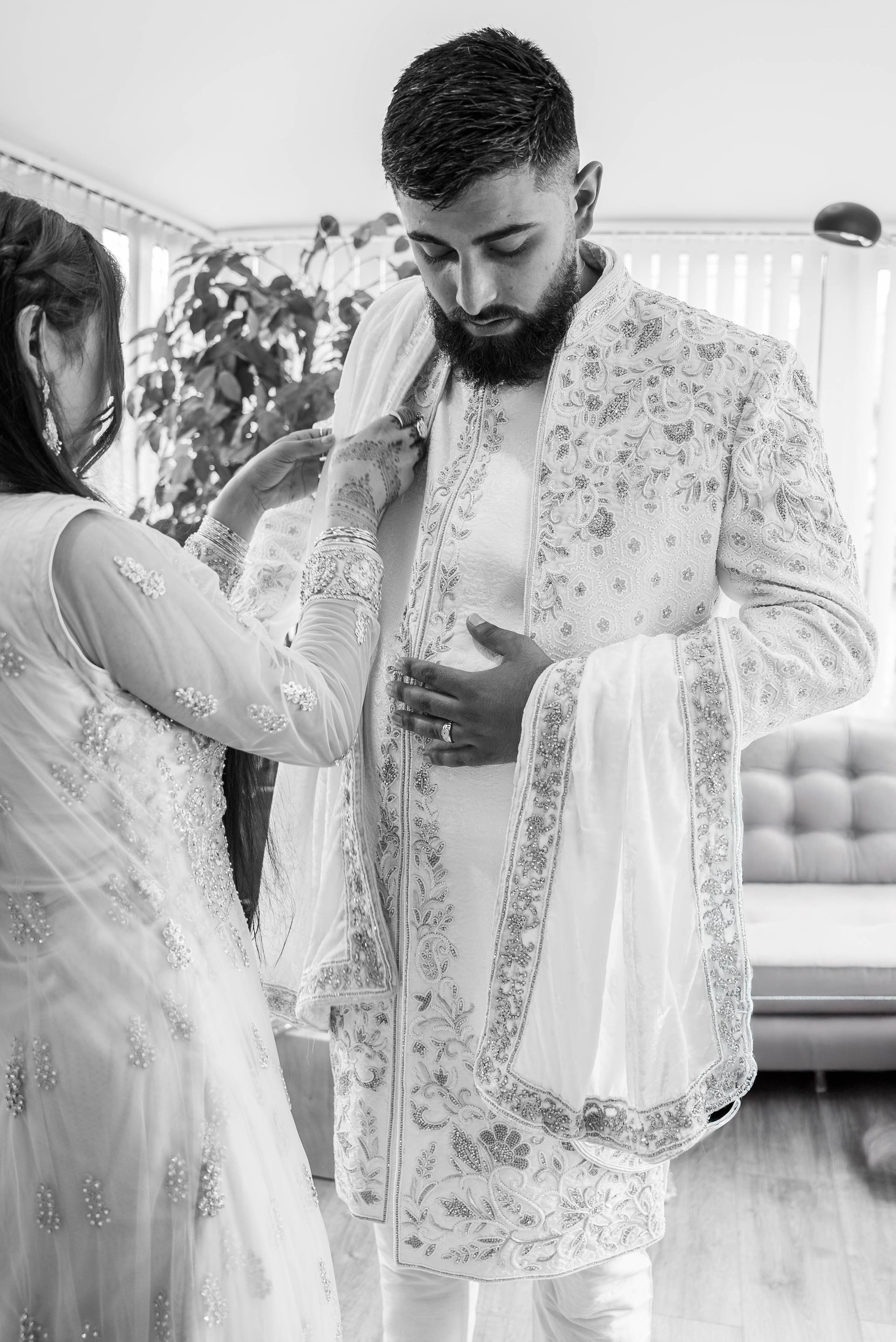 A man getting dressed in traditional, ornate attire, assisted by a woman in a richly embroidered dress, possibly during a cultural or religious ceremony.
