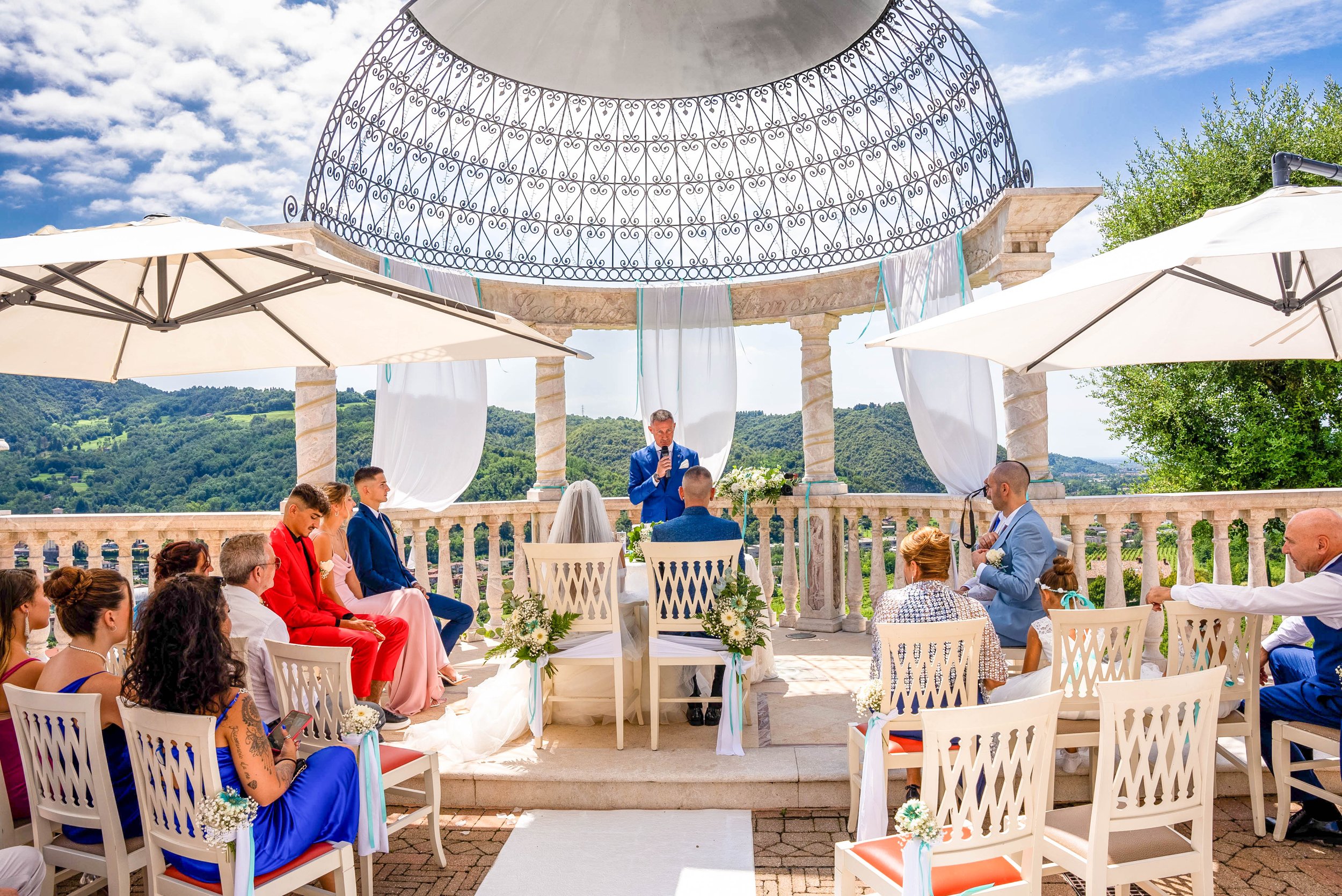 Wedding ceremony taking place outdoors on a terrace with hills in the background. The bride and groom are seated in front of a celebrant, with guests sitting at tables decorated with flowers around them. Large umbrellas provide shade, and an ornate m