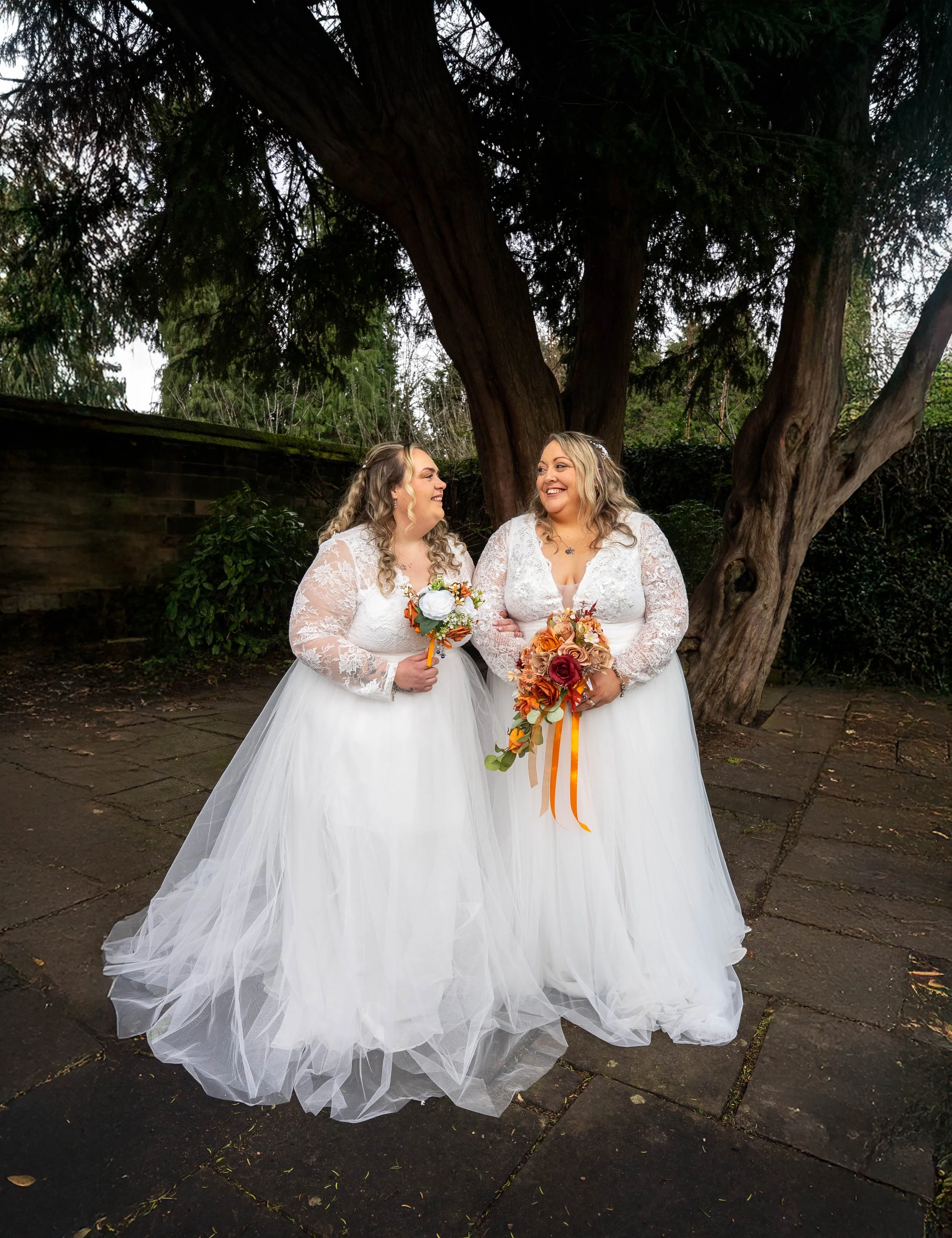 Two women in white wedding dresses standing outdoors under a large tree, smiling at each other, each holding a bouquet of flowers with autumn-colored roses and ribbons.