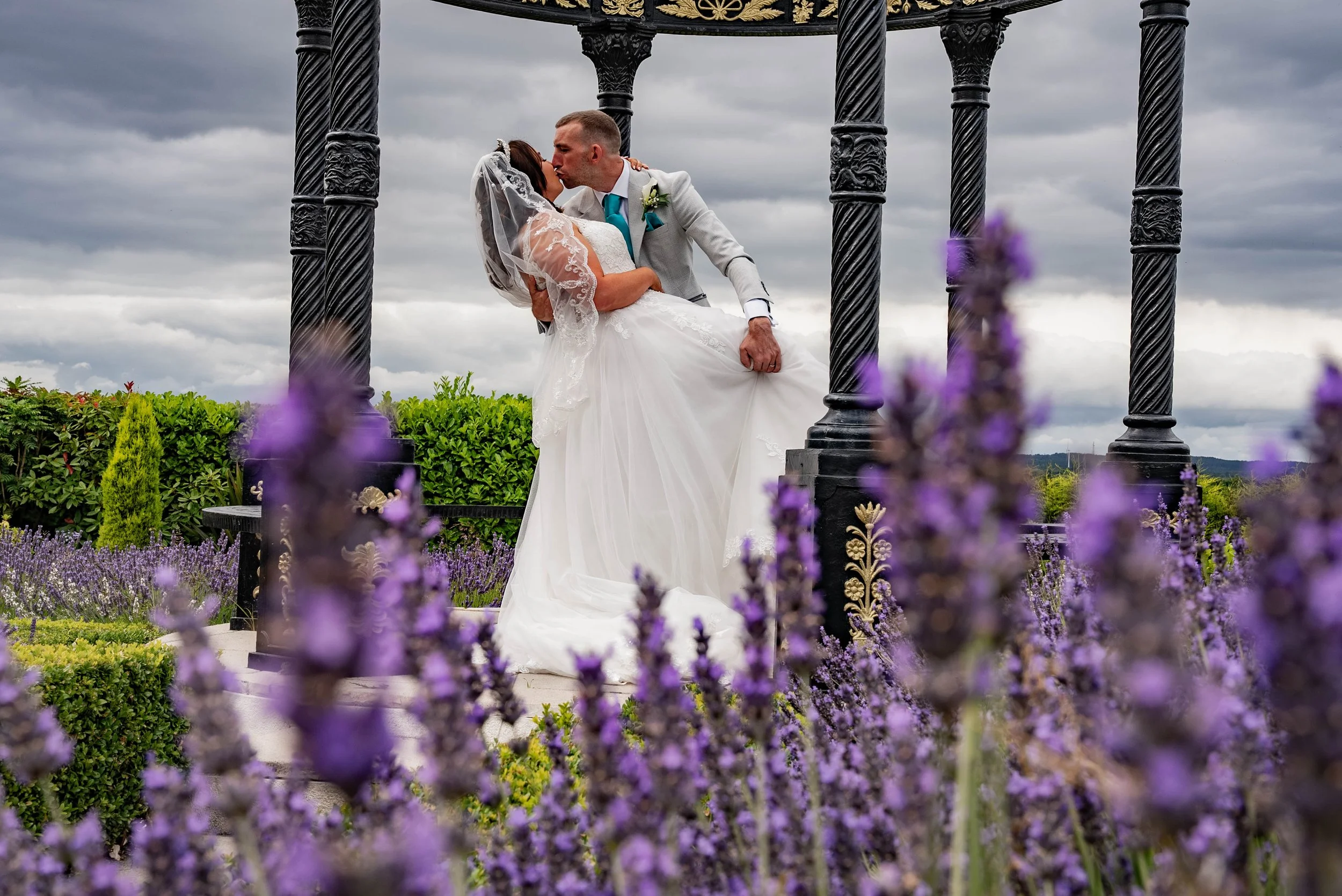 A newlywed couple sharing a kiss in an outdoor setting, with a black ornate gazebo and purple flowers in the foreground, under a cloudy sky.