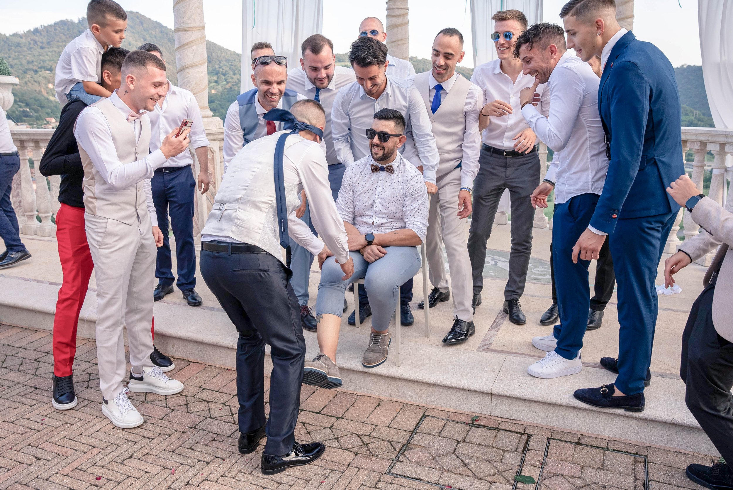 A group of men dressed in formal attire gathered outdoors on a terrace, engaging in a fun activity where one man is blindfolded and others are smiling and watching. The scene appears to be celebratory, with scenic mountains in the background.