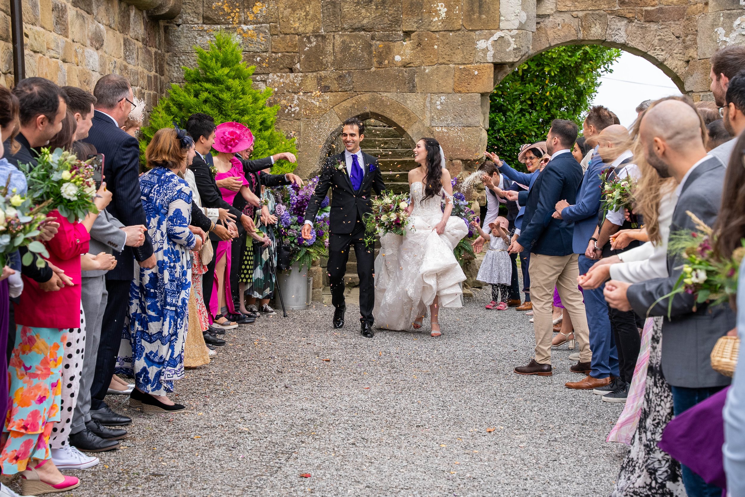 A newlywed couple walking through a crowd of people during their wedding celebration outside a stone building.