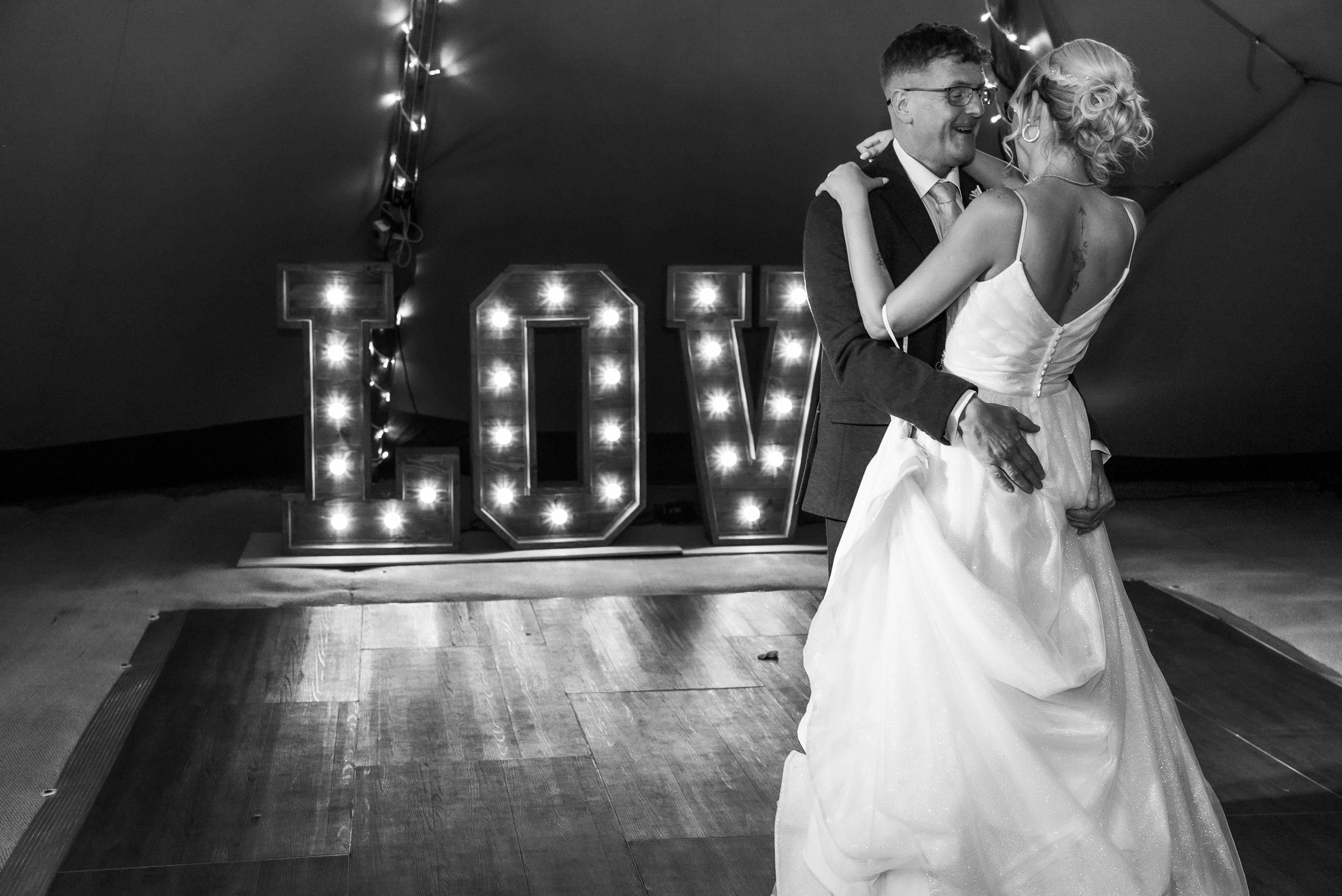 A bride and groom dancing at their wedding reception with large illuminated letters spelling 'LOVE' in the background.