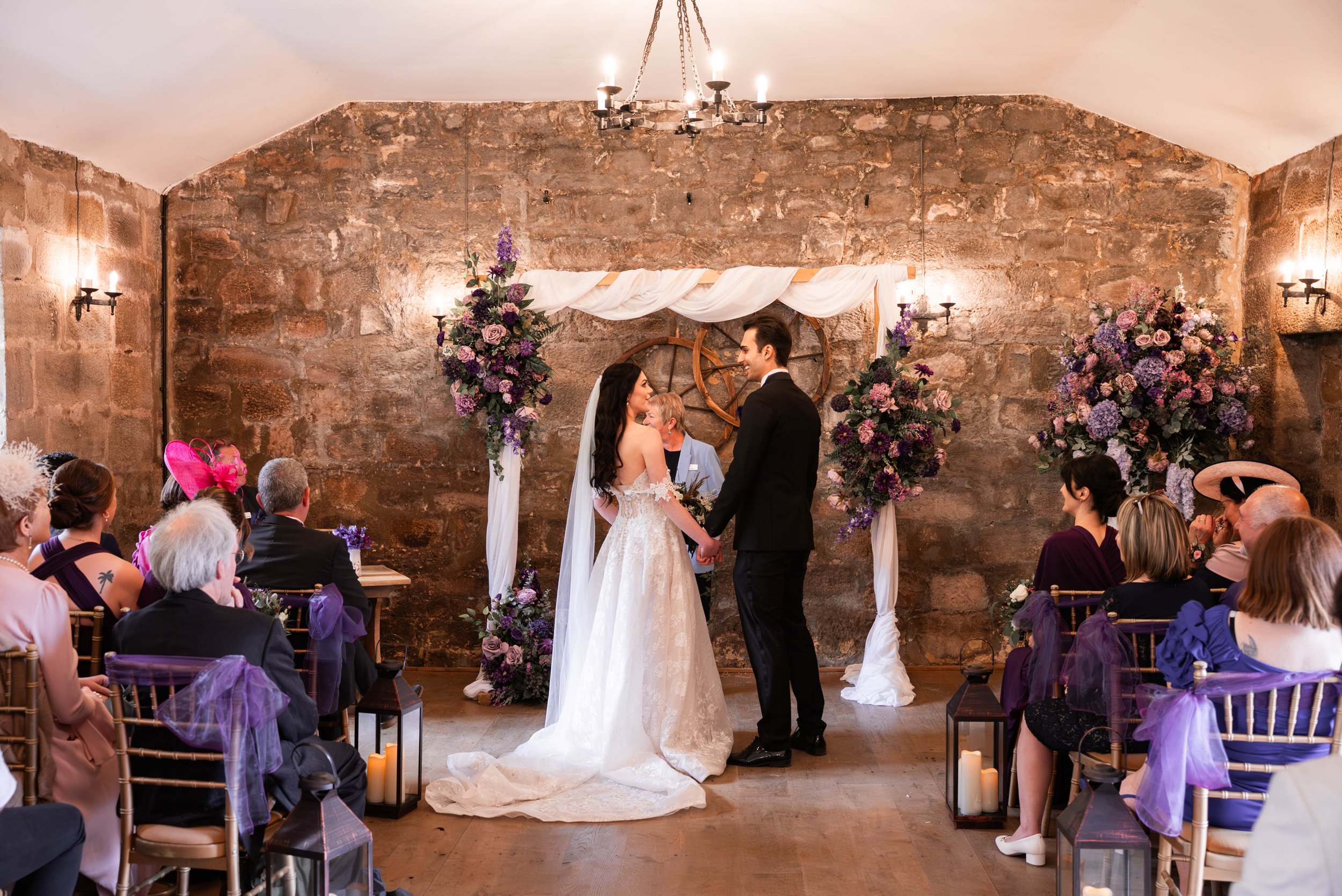 A wedding ceremony with a bride and groom holding hands, standing under a decorated arch with purple and pink flowers, in a rustic indoor venue with brick walls and hanging lanterns, with guests seated on either side.