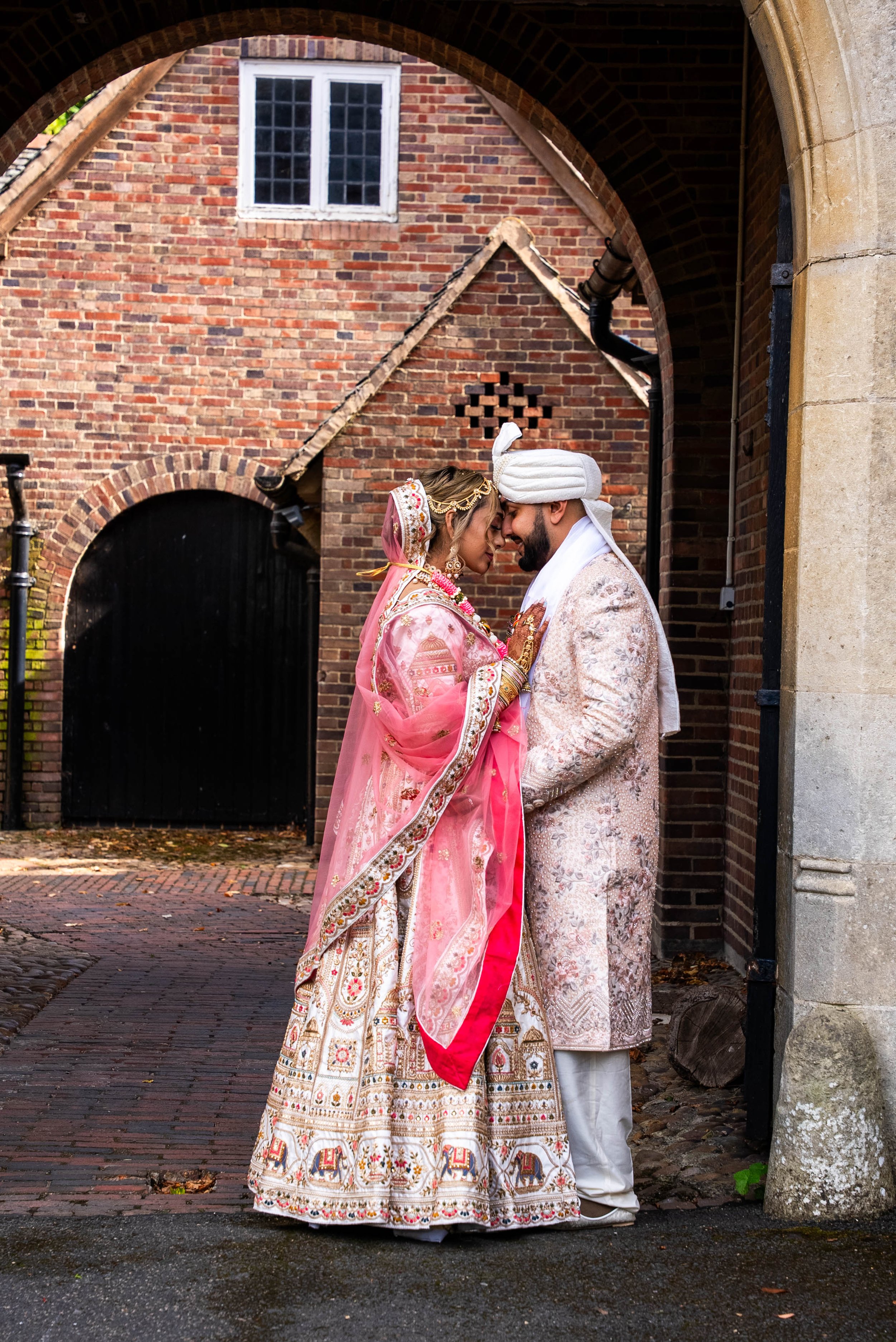 A bride and groom in traditional Indian wedding attire standing close together and touching foreheads in front of a brick building archway.