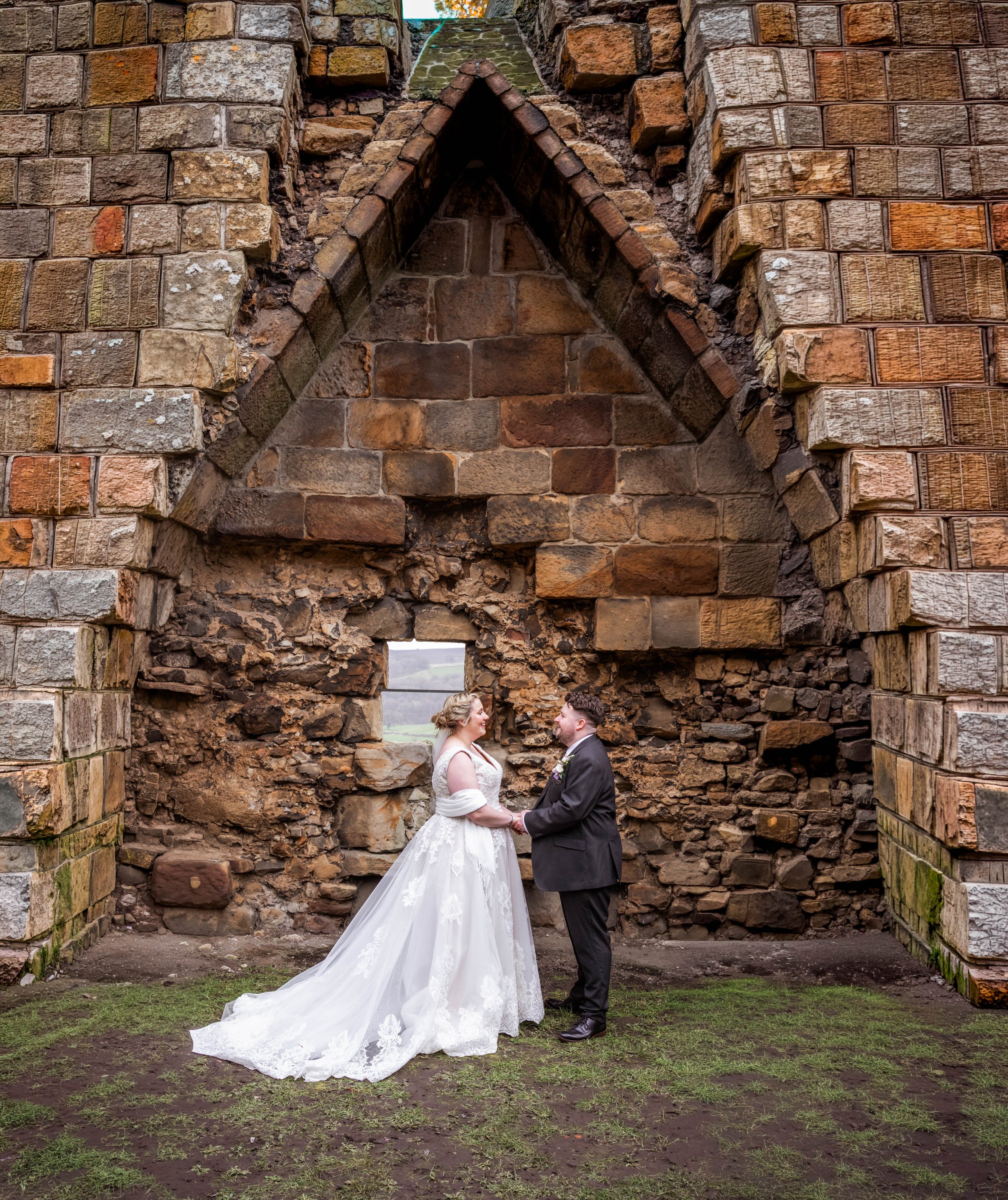 A bride and groom holding hands, standing face-to-face in front of a rustic stone structure with a triangular window and an arched doorway, outdoors on a grassy area.