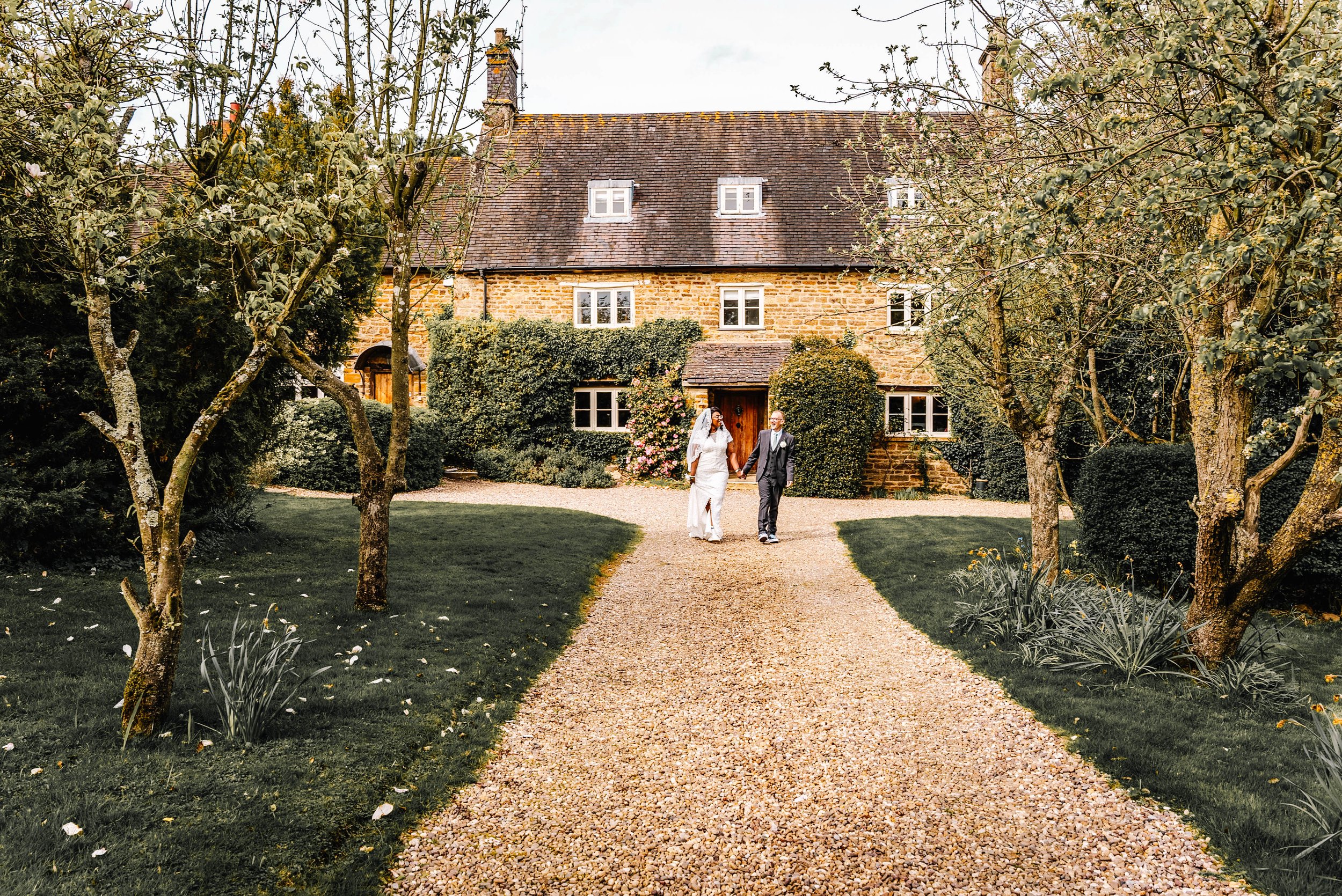 A bride and groom walking hand in hand on a gravel pathway leading to a brick house with a garden surrounded by trees and shrubs.