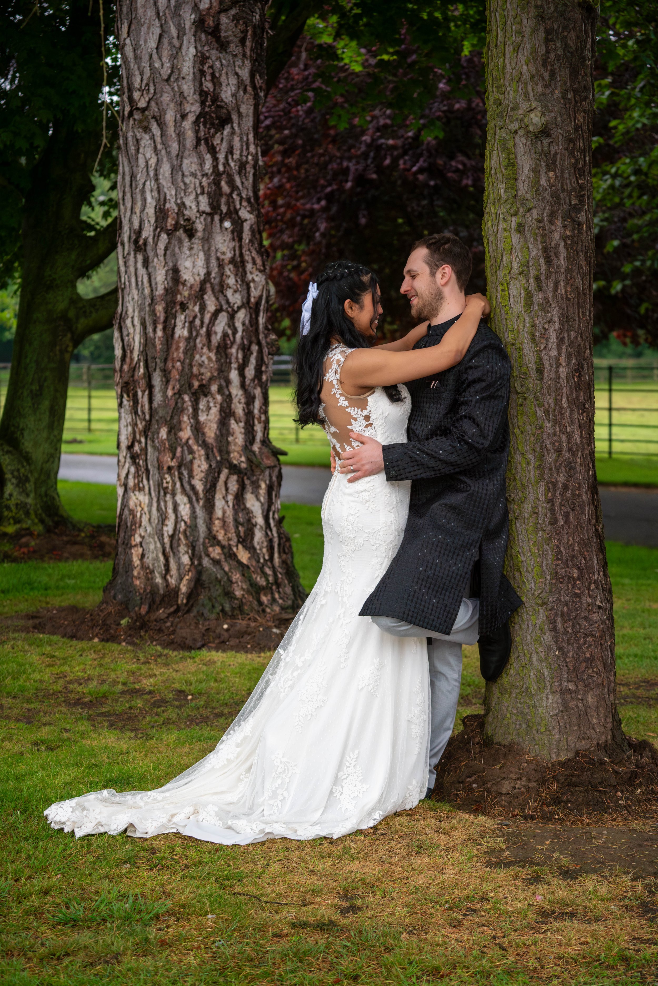 A bride and groom are embracing and smiling, standing between two large trees outdoors on a grassy area during daytime.