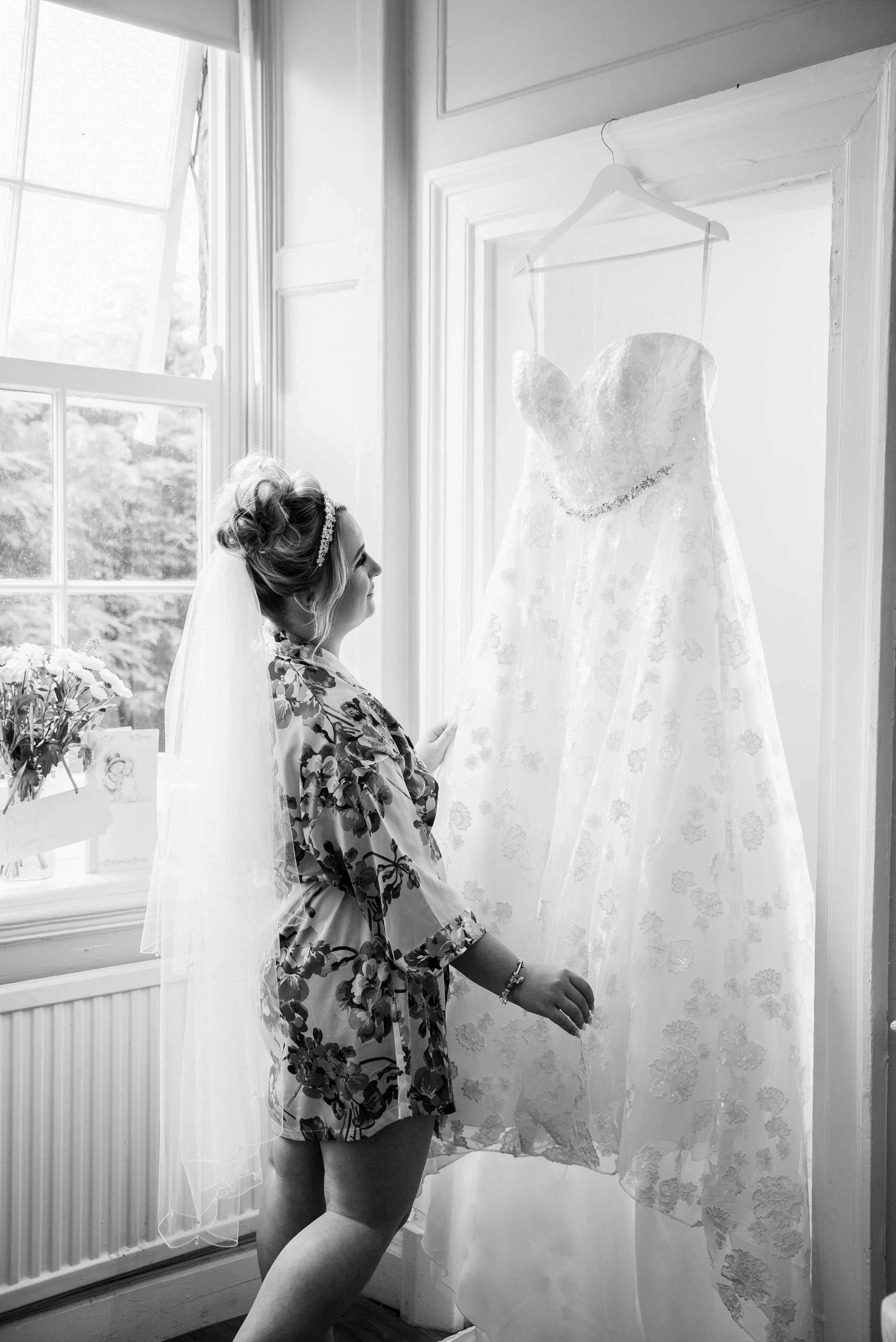 A bride-looking woman with a veil and floral robe gazing at a wedding dress hanging on a wall near a window.