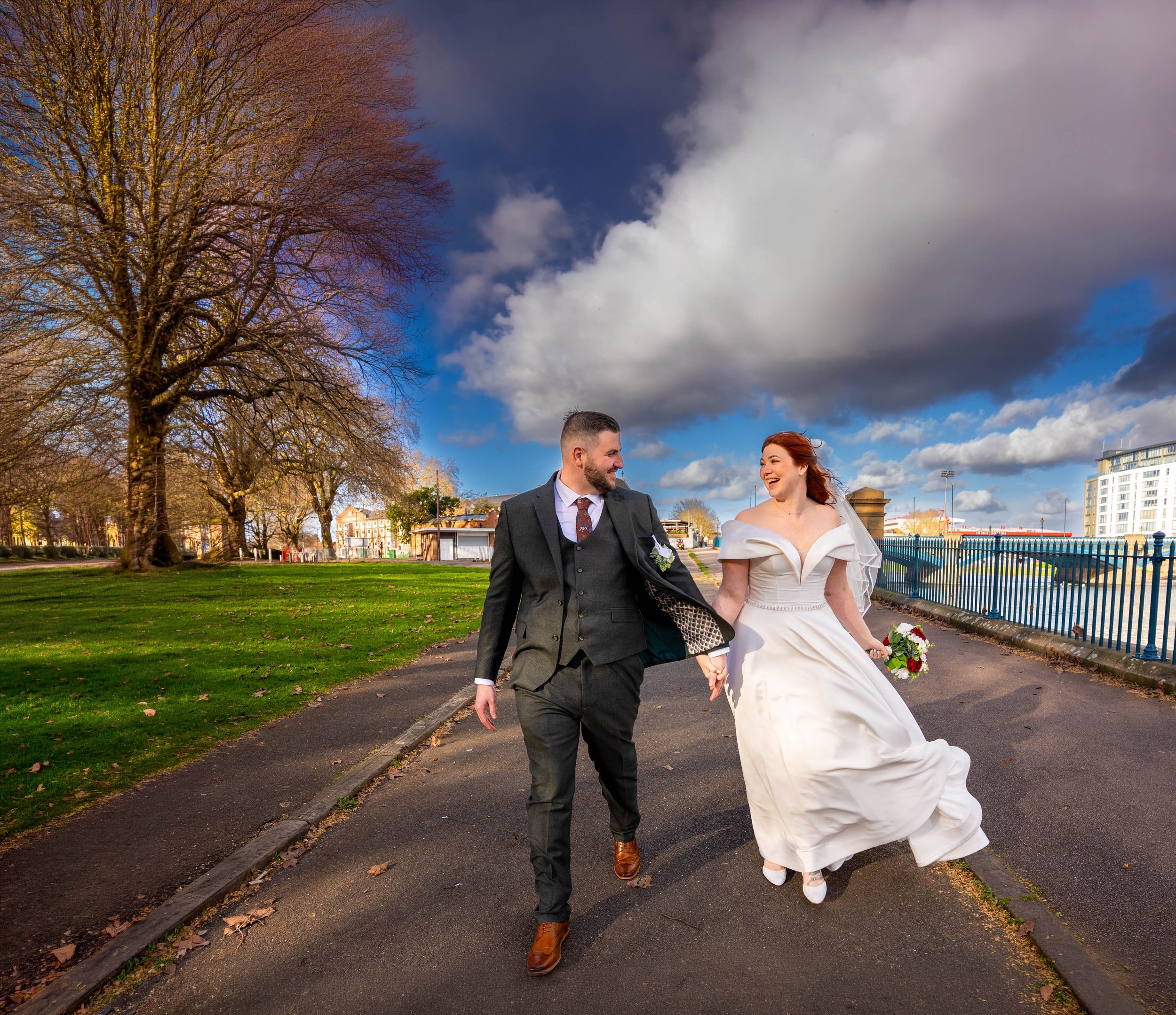 A smiling couple, dressed in wedding attire, walking hand-in-hand outdoors on a paved path in a park with green grass, leafless trees, and a cloudy sky.