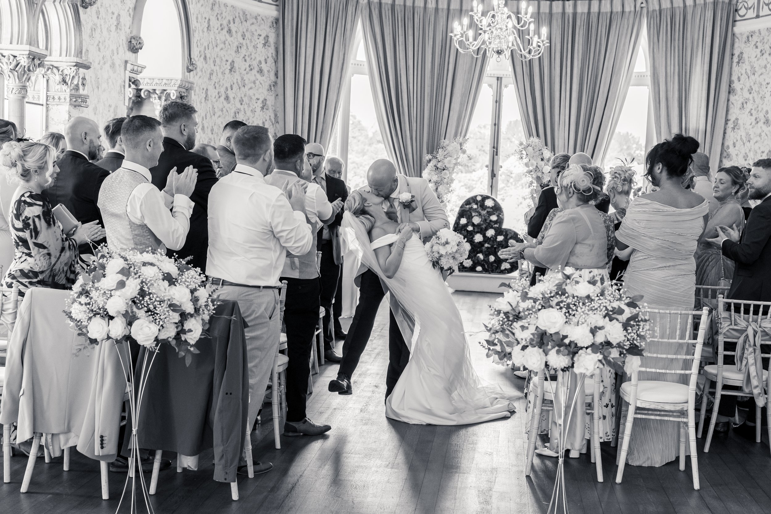A wedding ceremony with a bride and groom sharing a kiss as guests stand around them in a decorated elegant room with floral arrangements and large windows with curtains.
