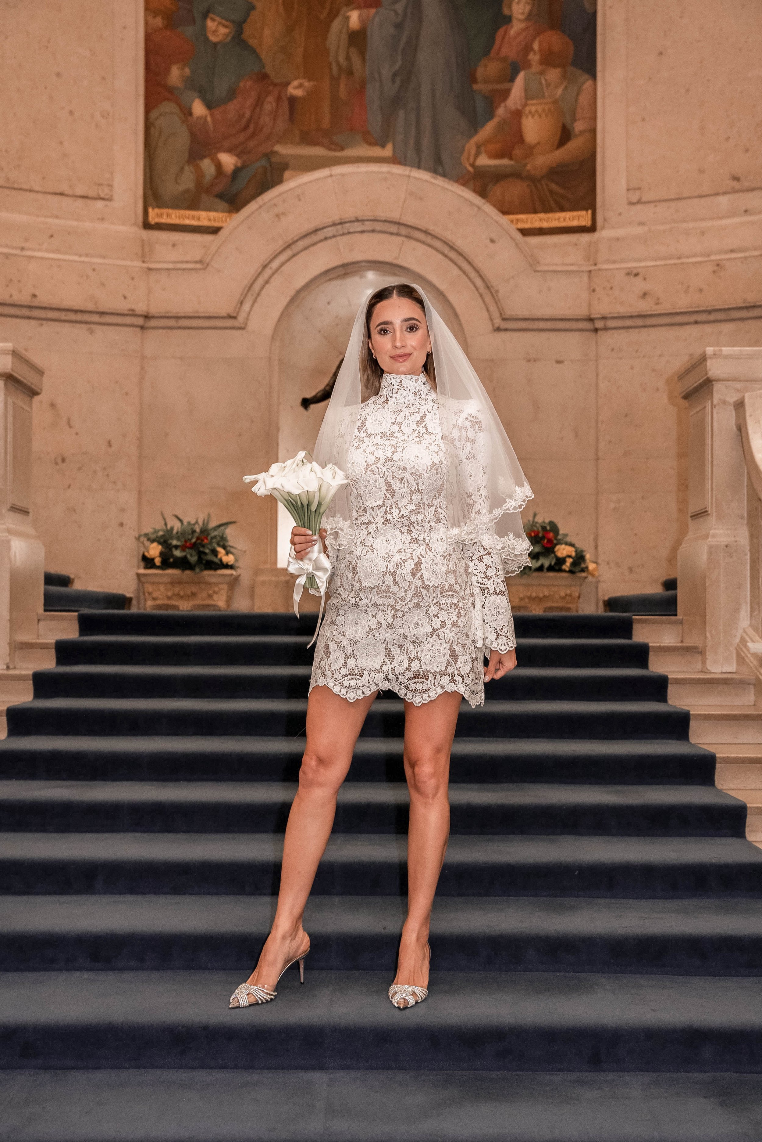 A woman in a lace wedding dress and veil holding a bouquet of calla lilies stands on a staircase inside a church or cathedral, with murals and floral arrangements in the background.