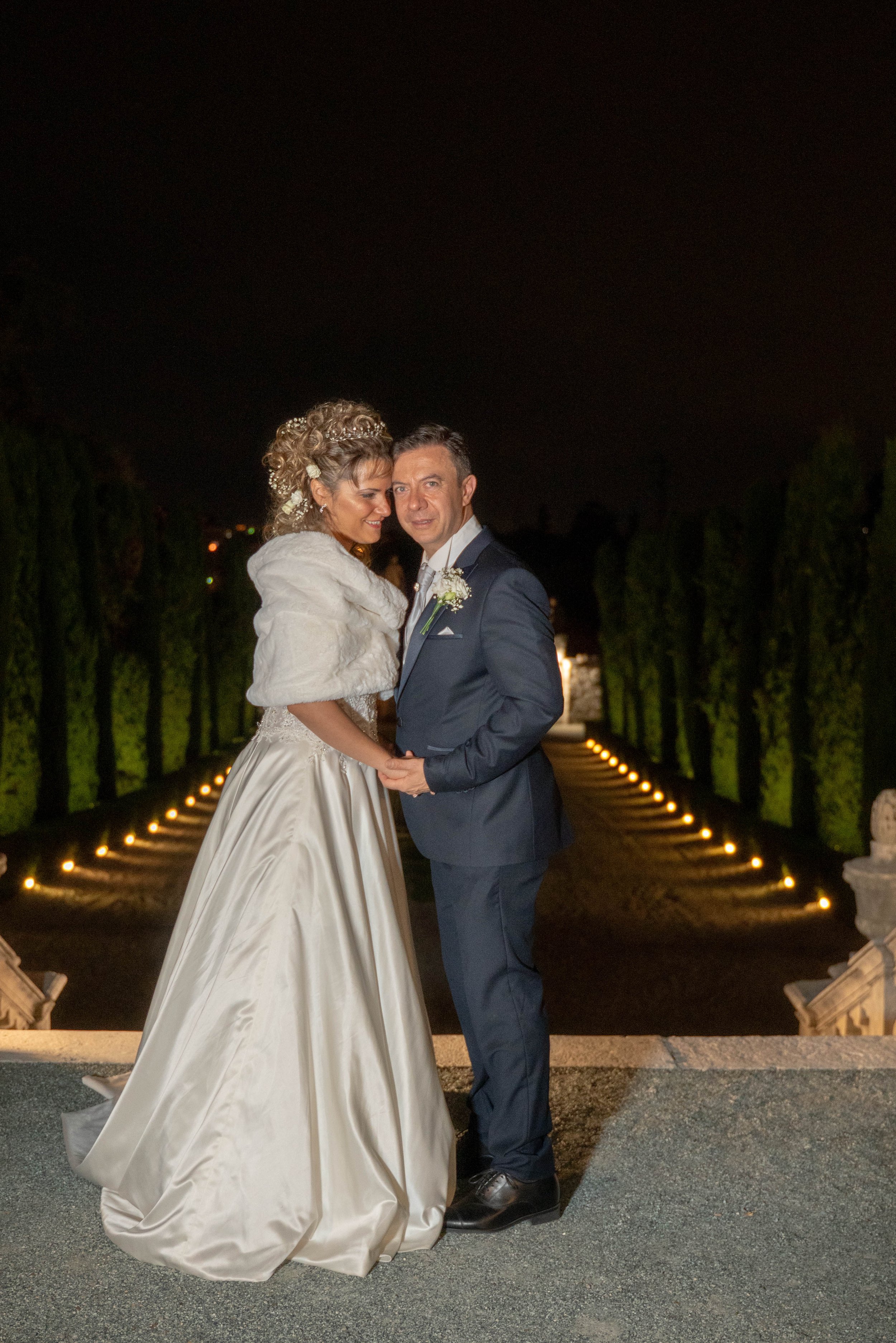 A bride and groom stand close together on an outdoor night, holding hands and smiling. The bride wears a white wedding gown with a faux fur shawl and has curly hair with floral embellishments. The groom wears a navy suit, white shirt, and tie, with a