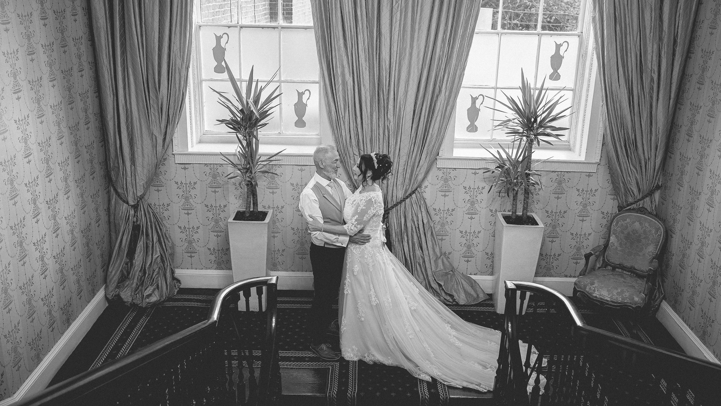 A bride and groom in wedding attire sharing a moment in a vintage-style room with patterned wallpaper, heavy drapes, and large windows decorated with vase silhouette decals.