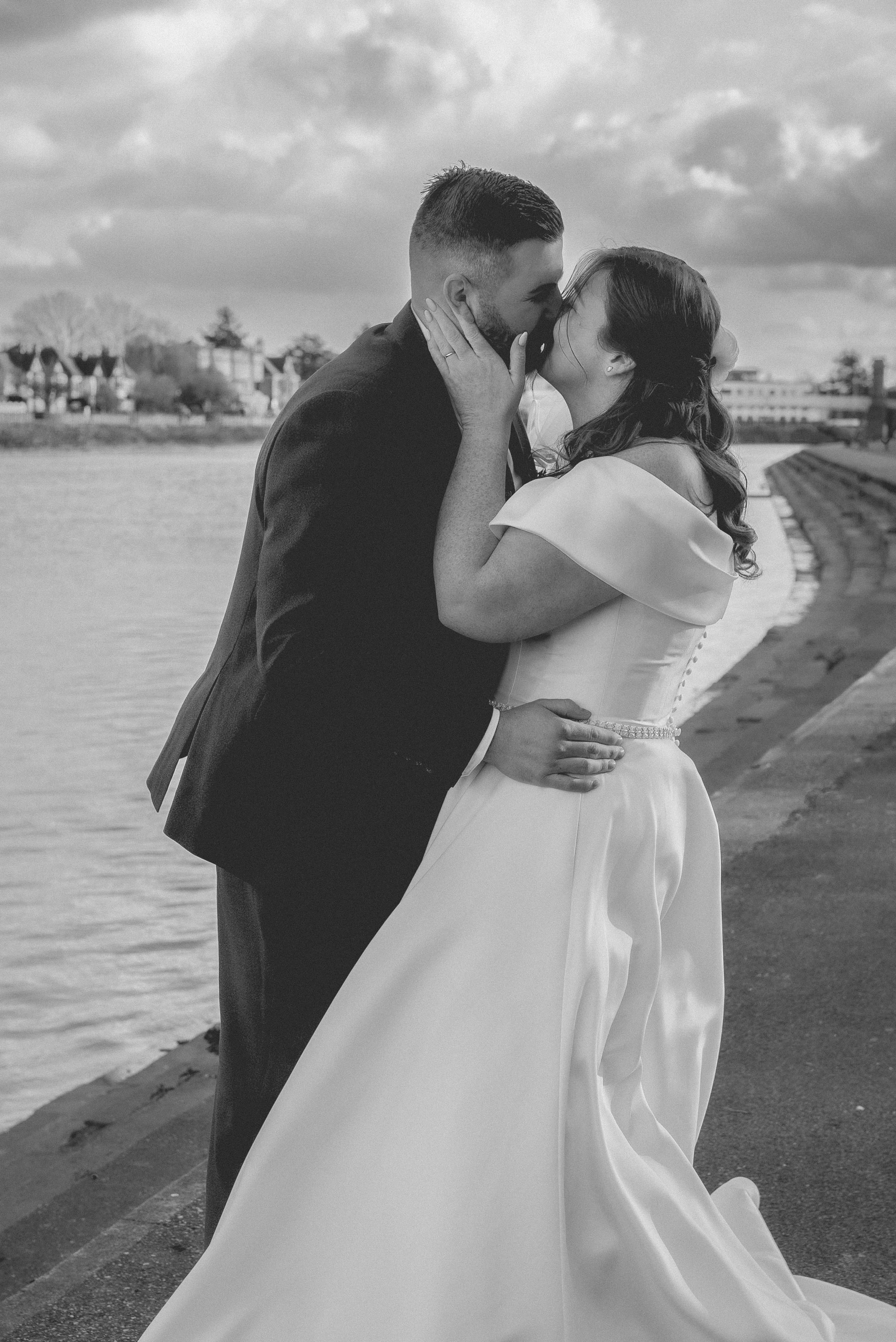 A black-and-white photo of a newlywed couple kissing by the water, with the bride in a flowing wedding dress and the groom in a dark suit, standing on a scenic waterfront pathway.