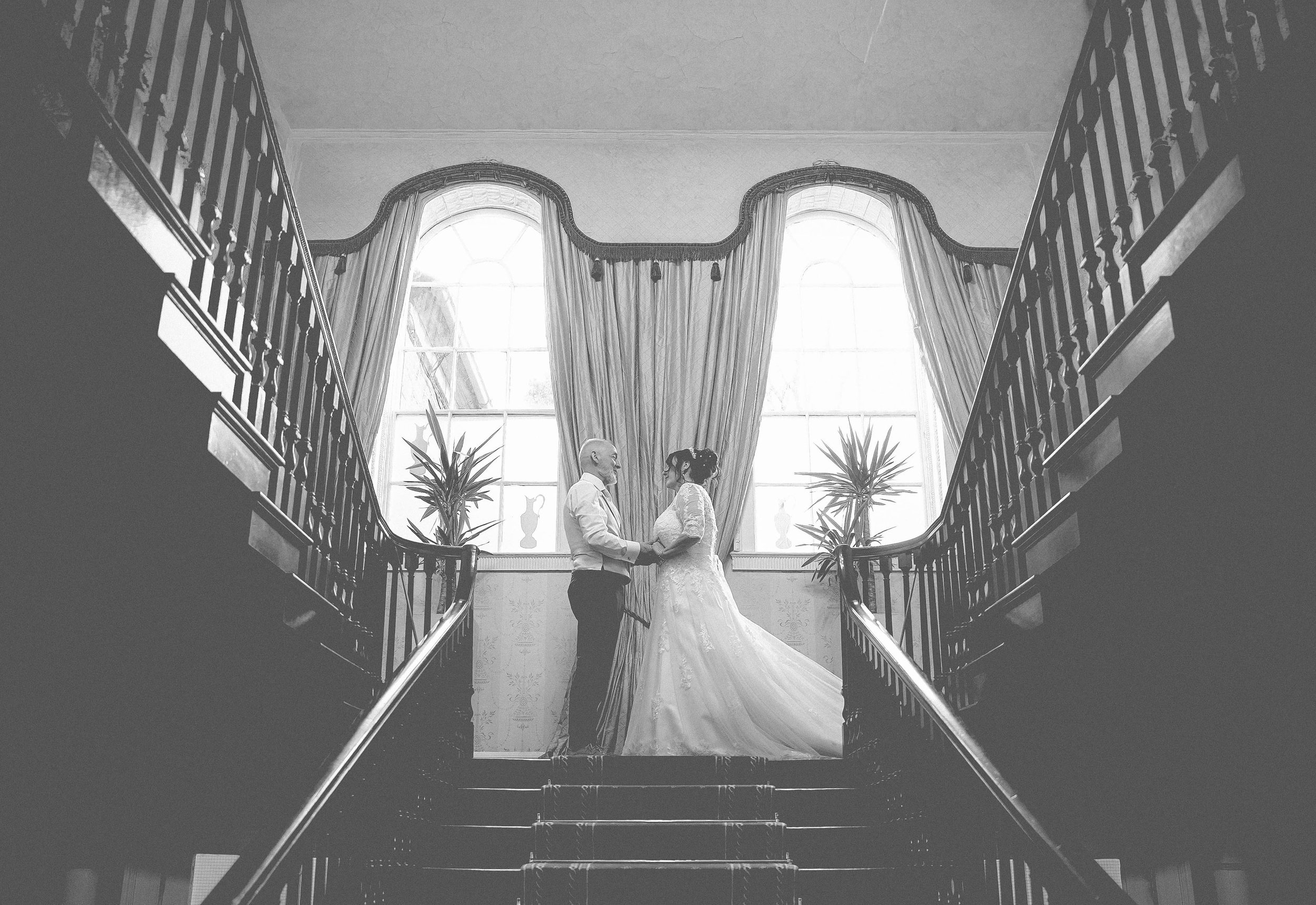 A bride and groom holding hands on a staircase in front of large windows with curtains and plants, during a wedding ceremony.