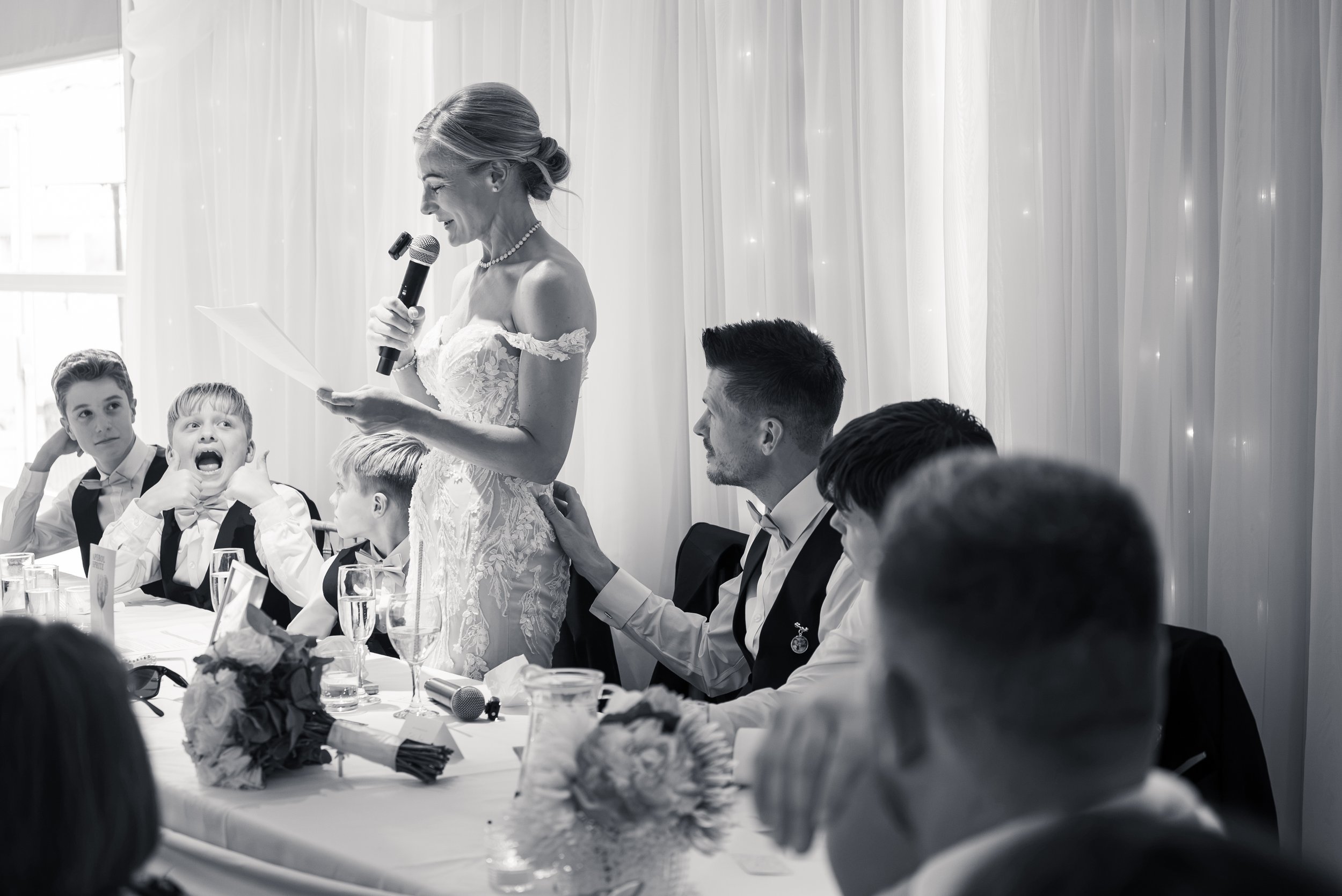 A woman in a wedding dress speaking into a microphone at a wedding reception with children and guests seated at a decorated table.