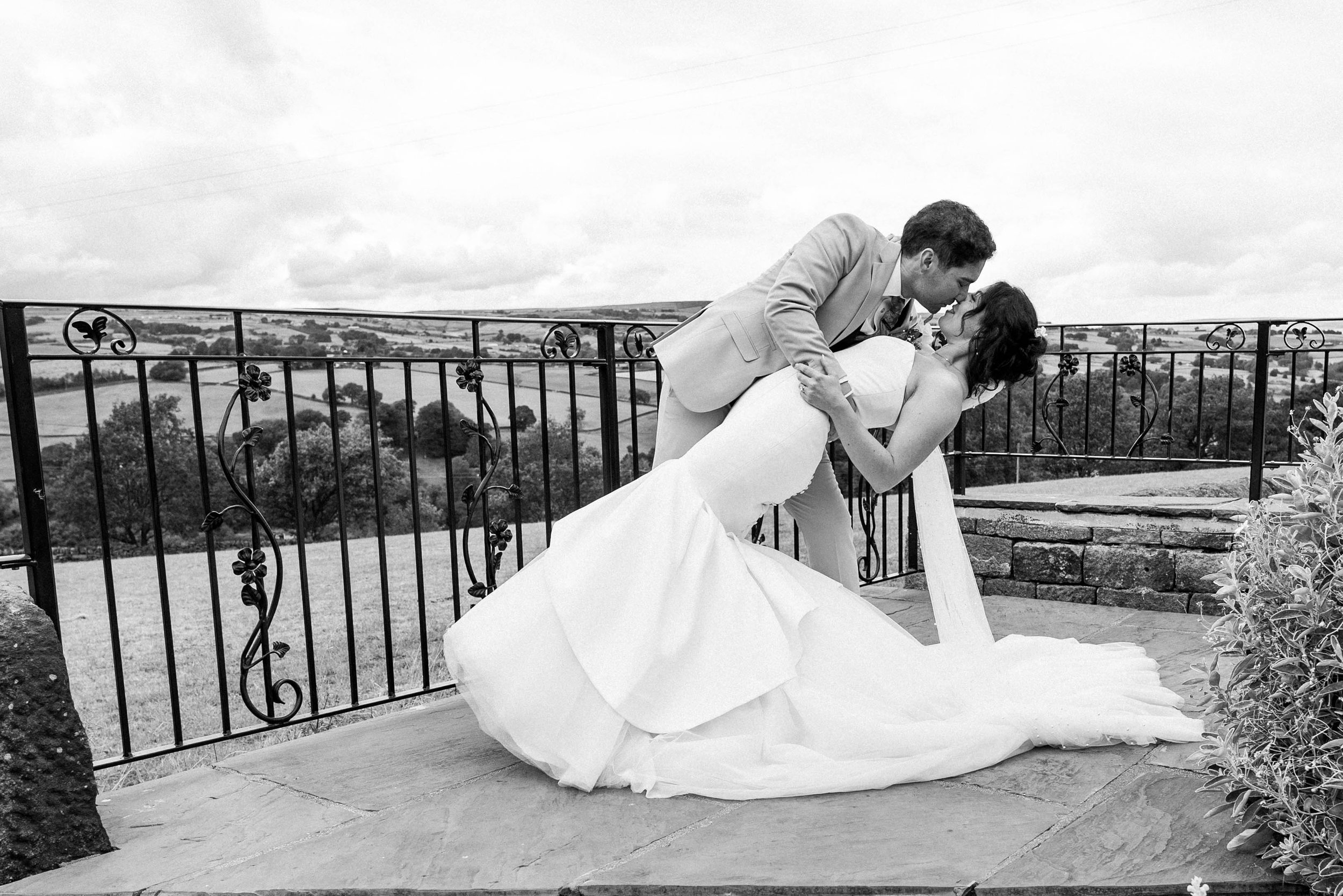 A black and white photo of a couple on a terrace with a scenic rural landscape in the background. The man, dressed in a suit, is dipping the woman, who is wearing a wedding gown, for a kiss.