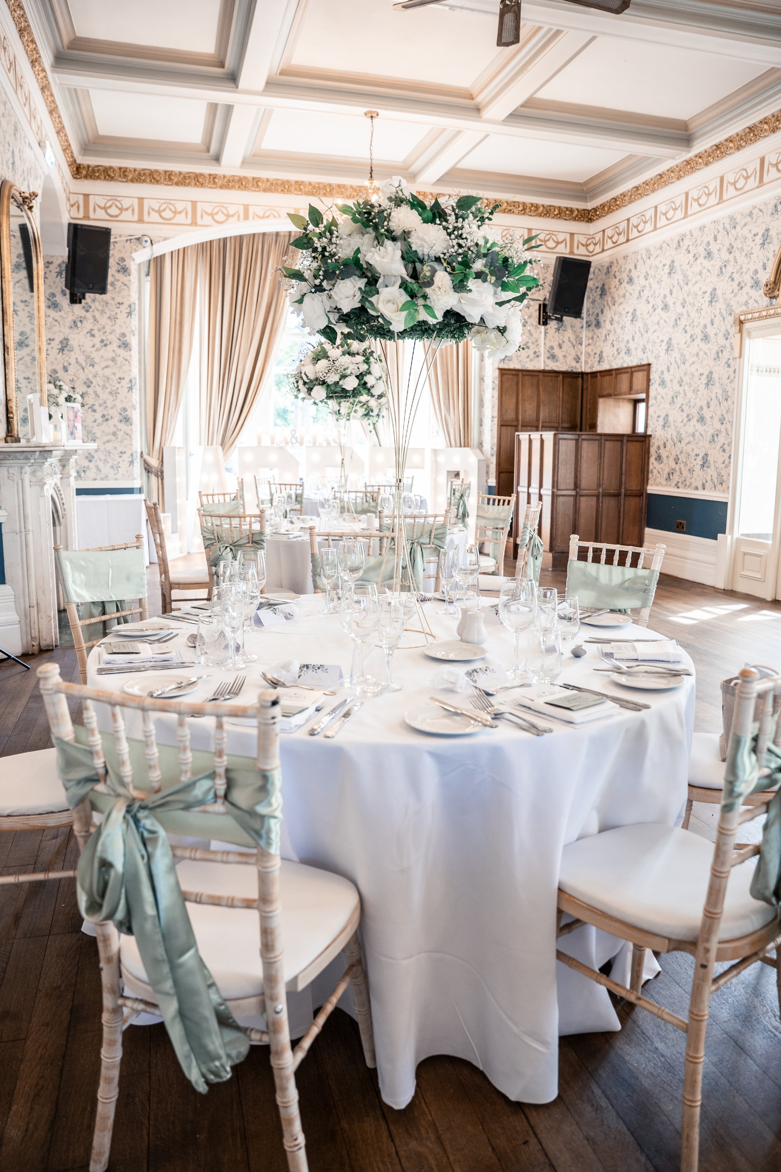 Elegant banquet room with round tables covered in white tablecloths, set with glassware, plates, and silverware. Tall floral centerpieces with white flowers and green foliage decorate each table. Wooden chairs with pastel green satin sashes tied in b