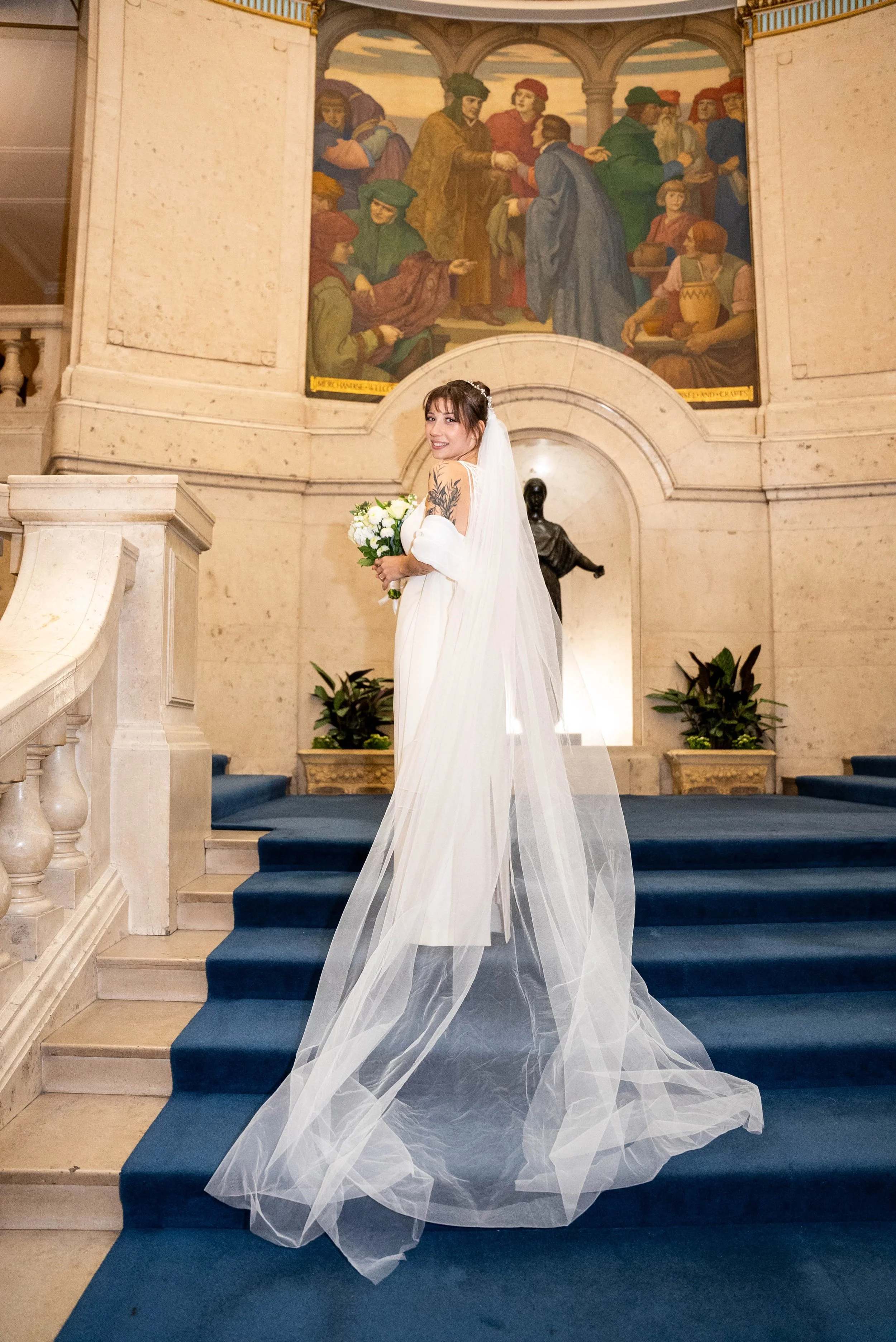 A bride in a white wedding dress with a long veil holding a bouquet of white flowers, standing on a staircase inside a grand building with a large mural painting on the wall behind her.