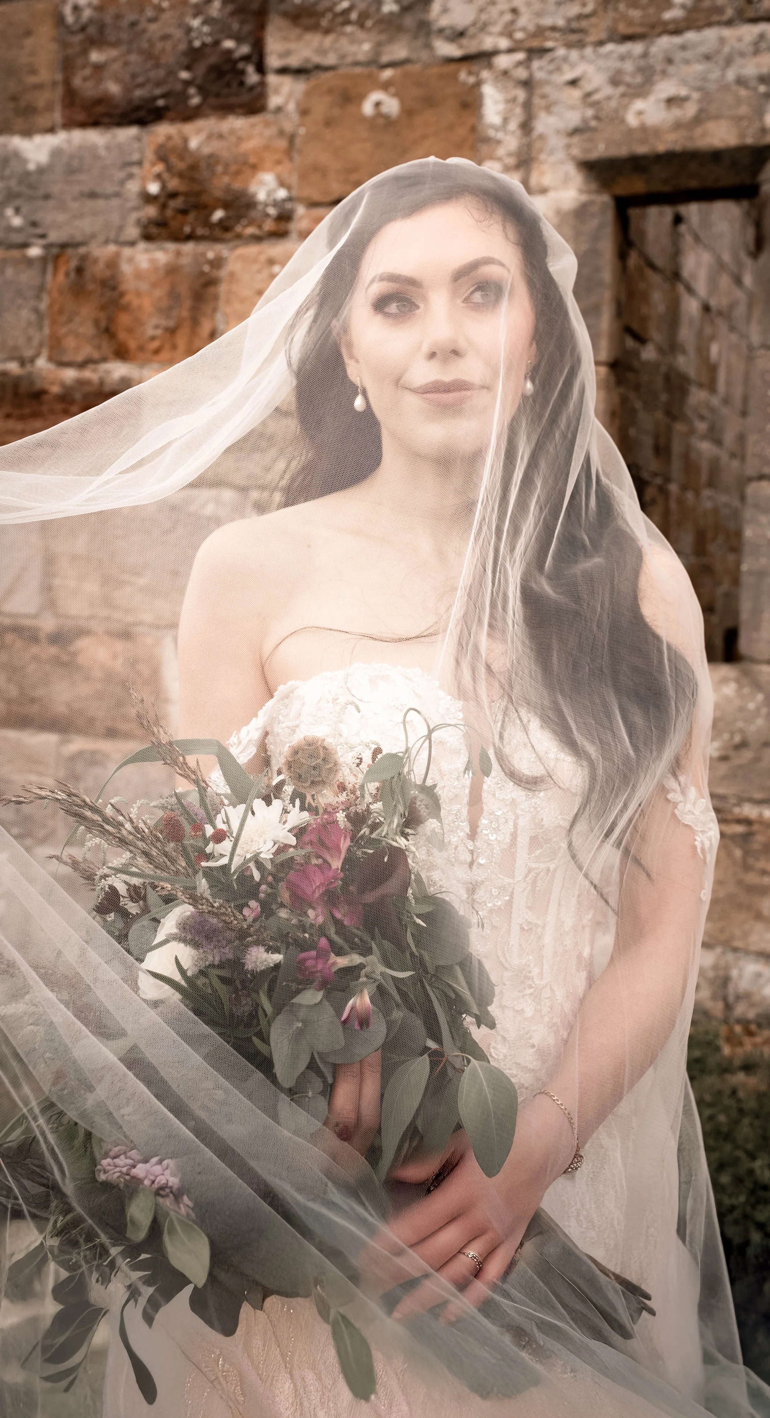 A bride in a white wedding dress holding a bouquet of flowers, with a veil over her face, standing in front of a brick wall.