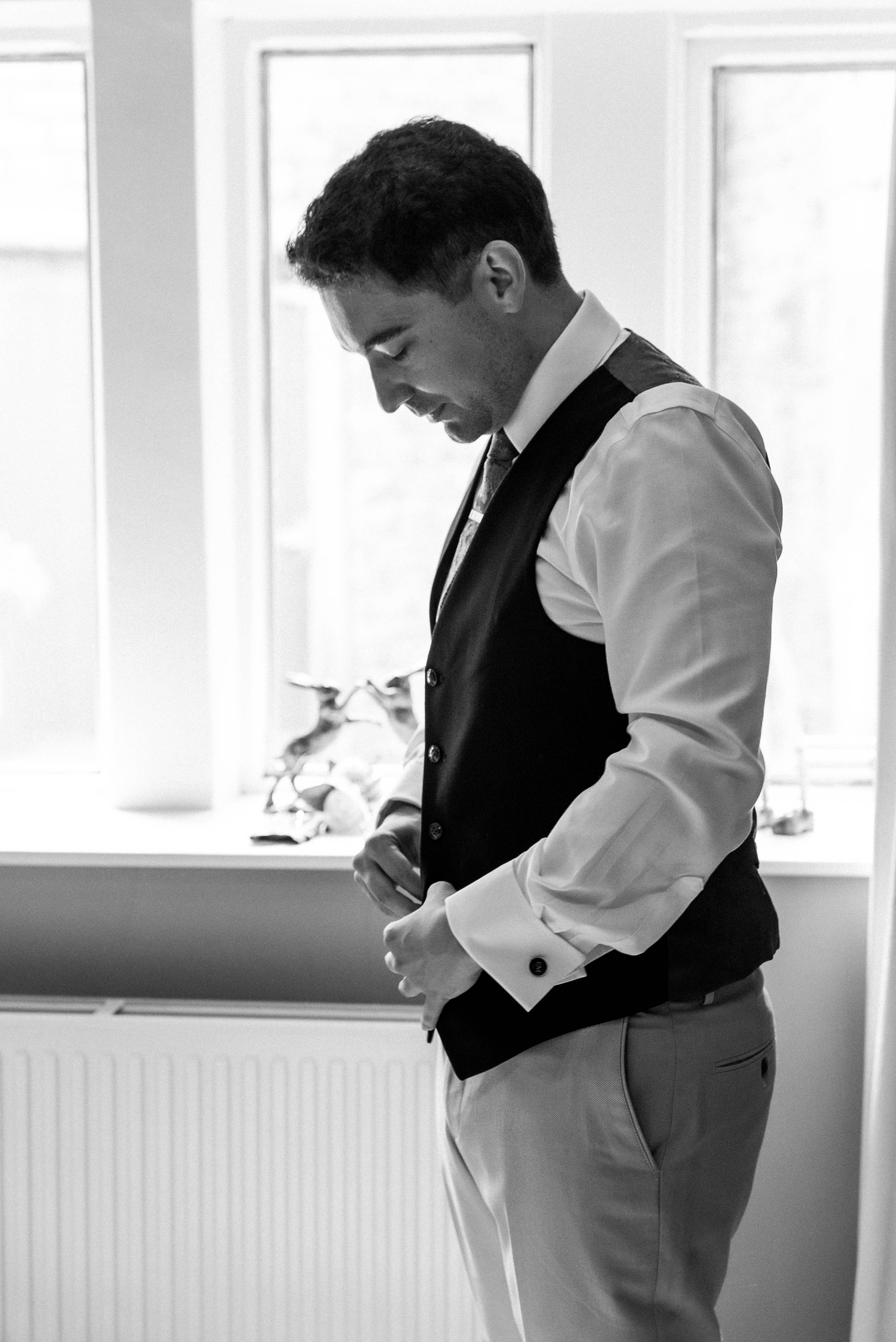 A man dressed in formal attire, including a vest, shirt, and tie, is preparing himself by adjusting his clothing, standing indoors near a window.